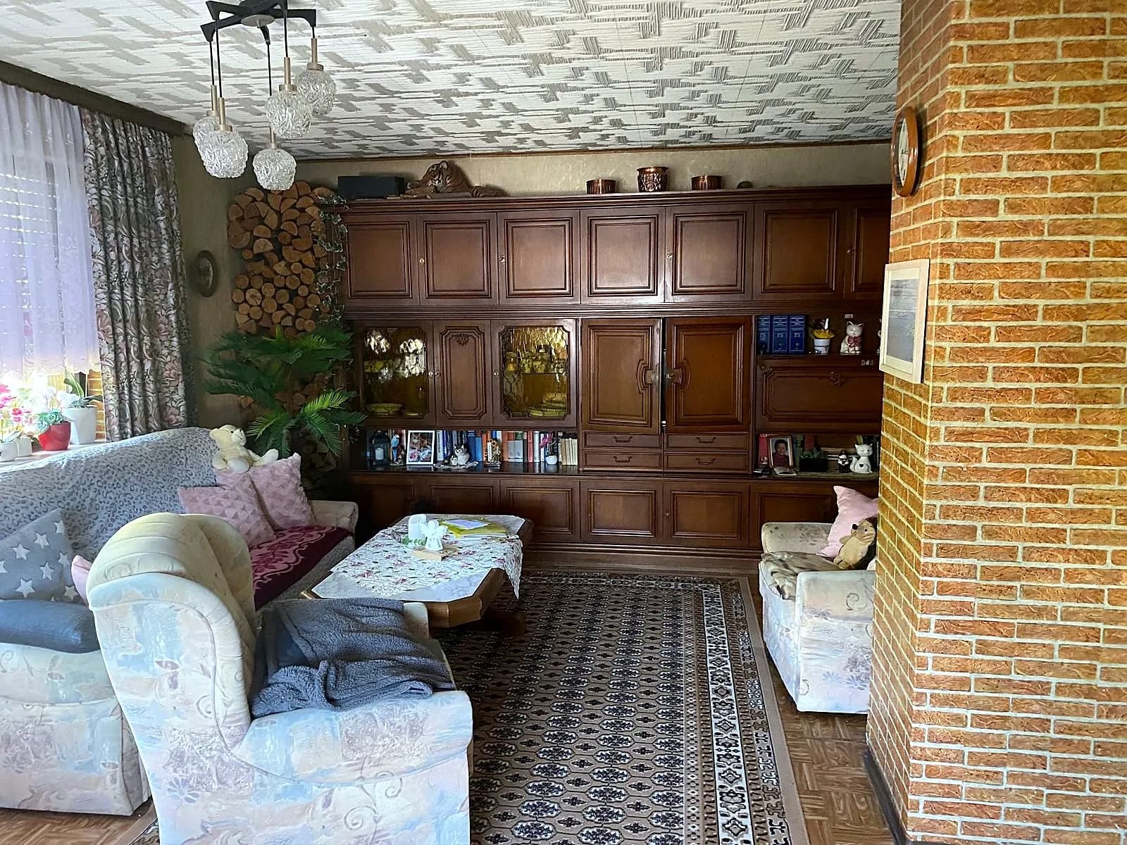 Gemütliches Wohnzimmer im traditionellen Stil mit rustikaler Holz-Schrankwand, bequemer Polstergarnitur, Couchtisch, Ziegelstein-Akzentwand und Kaminholzlager.