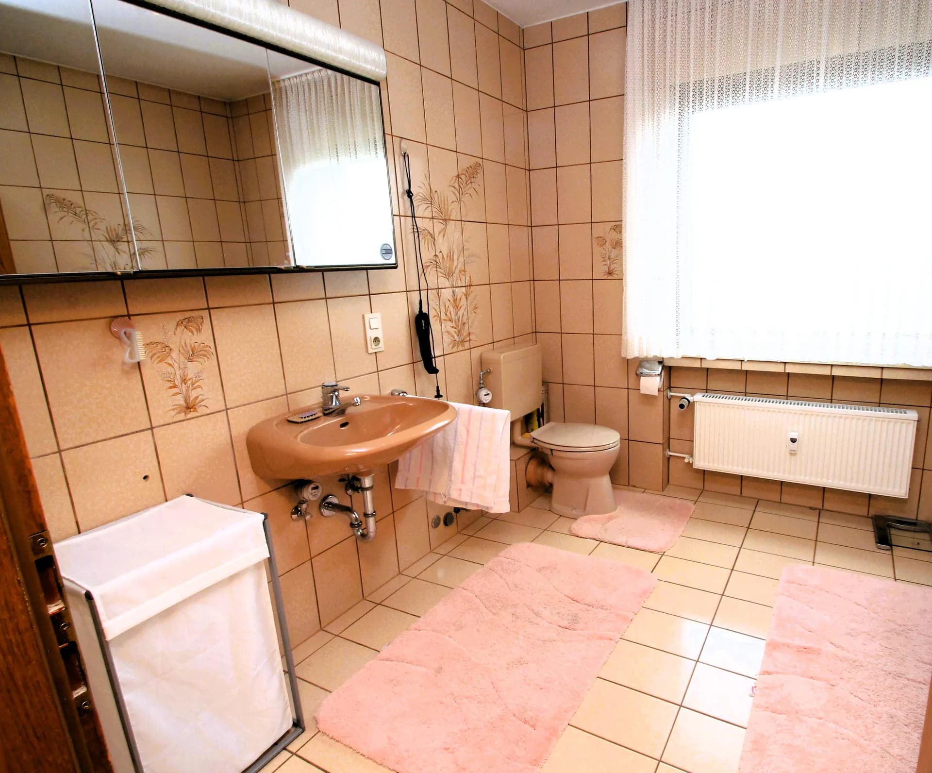 Großzügiges Wellness-Badezimmer mit beiger Verfliesung, Waschtisch, WC, antiker Holzkommode am Fenster, prachtvollem Kristallleuchter, vergoldetem Zierspiegel und königsblauem Samtsessel.