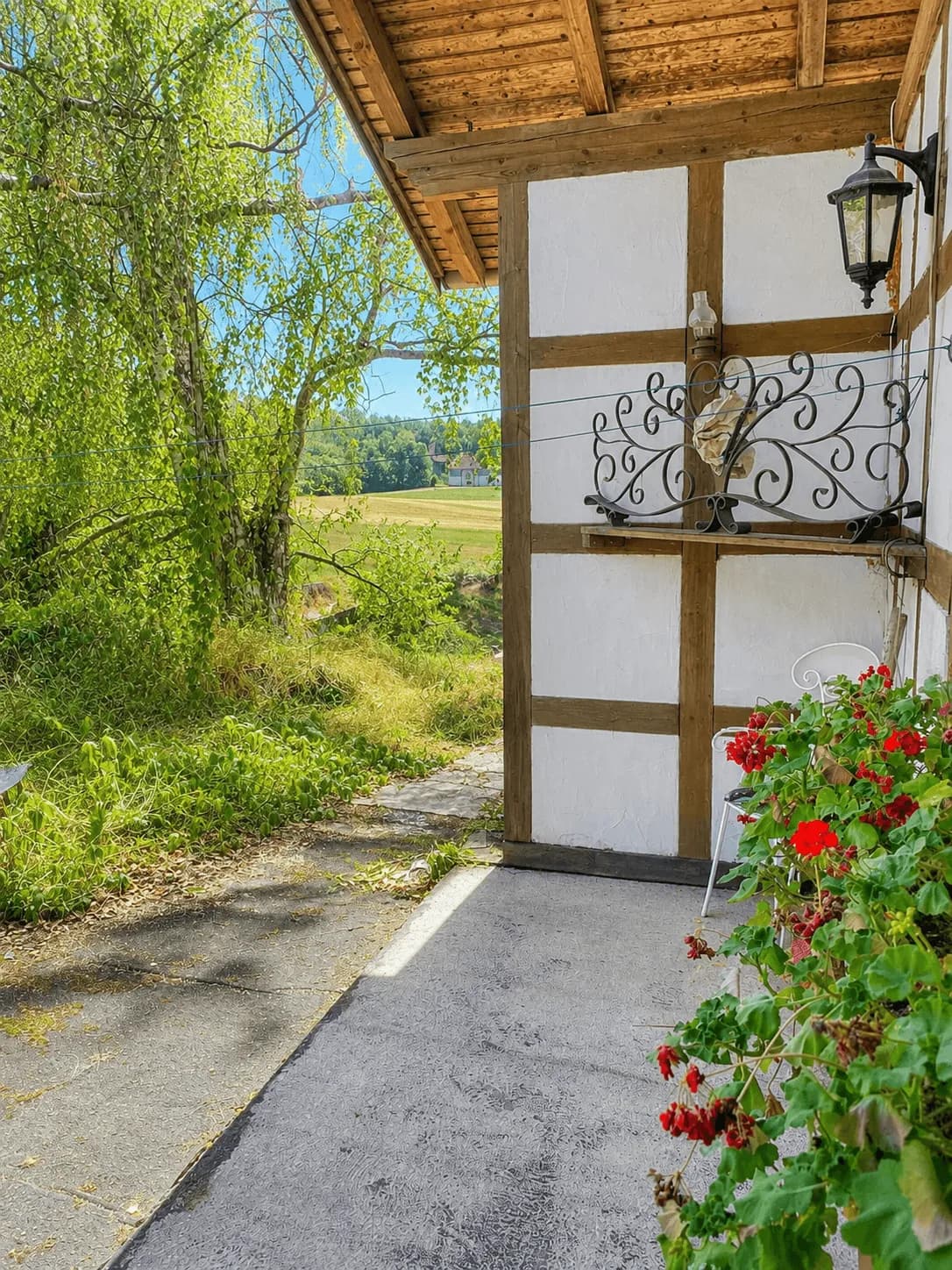 Überdachter Terrassenbereich mit Fachwerkelementen, Zierschmiedekunst, Blumenbepflanzung und weitem Blick ins Grüne.