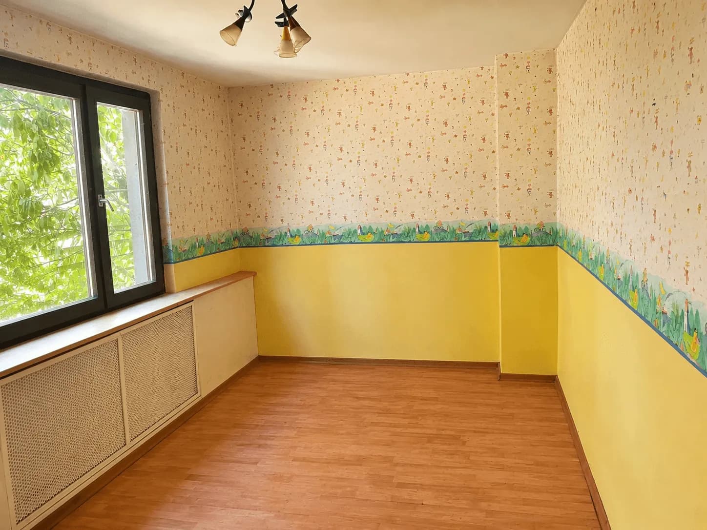 Helles Zimmer (Kinder- oder Arbeitszimmer) mit warmem Laminatboden, verkleidetem Heizkörper und Fenster mit Blick in den Garten.