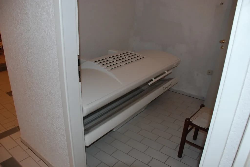 Separater Wellness- oder Ruhebereich mit professionellem Liegesolarium, hellem Fliesenboden und praktischem Holzhocker.