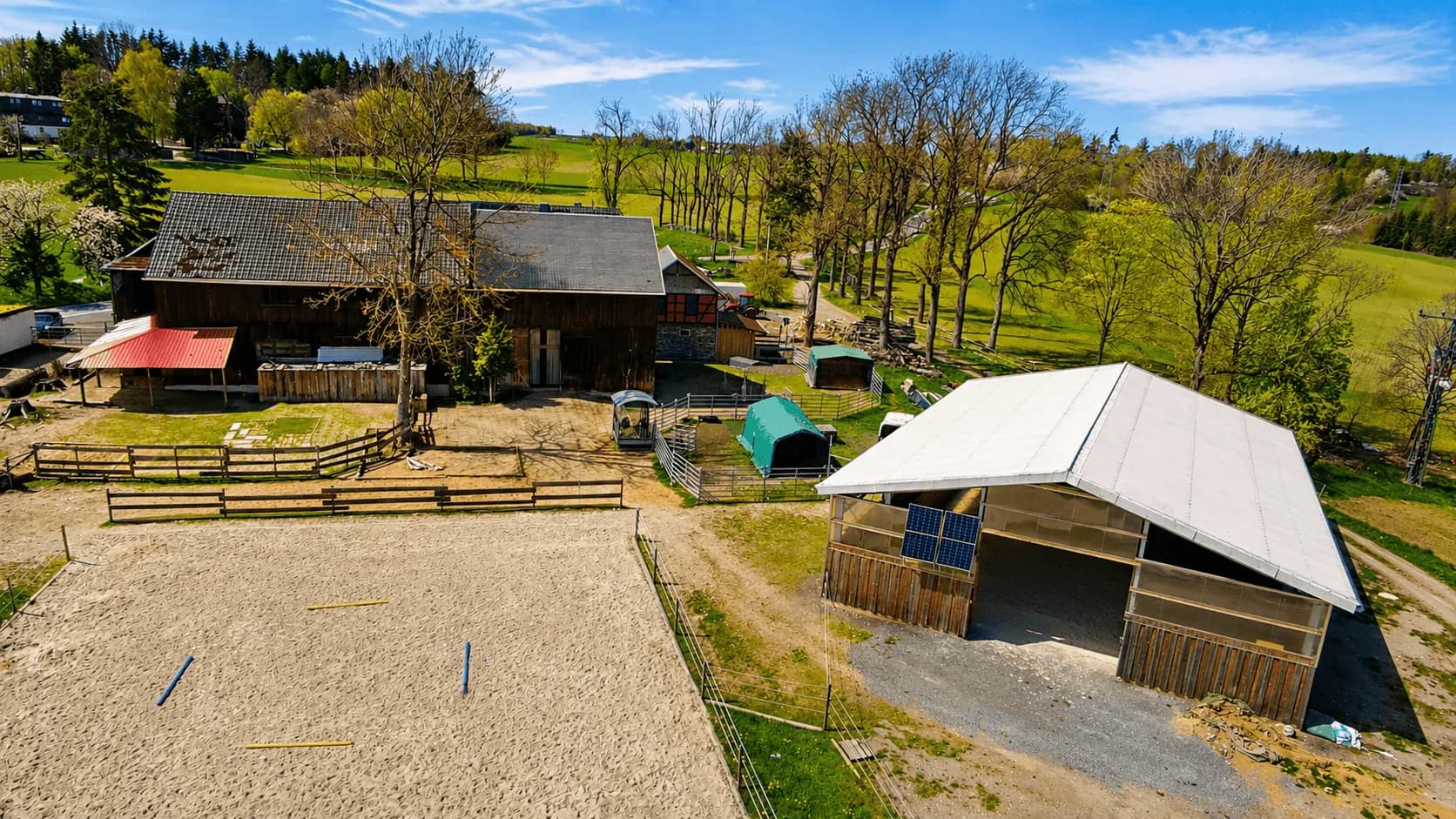 Luftaufnahme der Reitanlage/des Reiterhofs: Sandreitplatz mit Bodenstangen, offene Reithalle/Unterstand mit Solarmodul, traditionelle Holzscheune und weitläufige Weideflächen in ländlicher Umgebung.