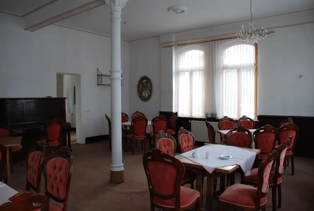 Traditioneller Gast- oder Speisesaal mit gepolsterten Barockstil-Stühlen, eingedeckten Tischen, weißer Ziersäule, Klavier und funkelndem Kronleuchter.