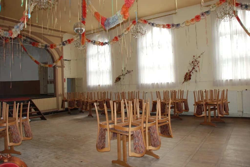 Großer Veranstaltungssaal mit bühnenartiger Nische, Echtholzparkett, hohen Fenstern mit Vorhängen, Diskokugel und für Festlichkeiten vorbereiteter Bestuhlung.