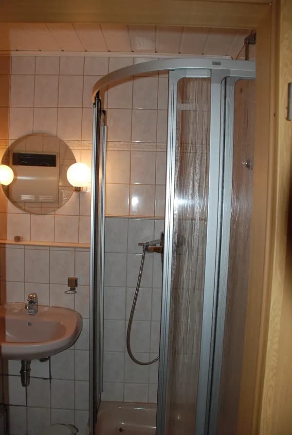Kompaktes Duschbad mit Runddusche, Waschbecken, rundem Wandspiegel samt Kugelleuchten und heller Verfliesung.