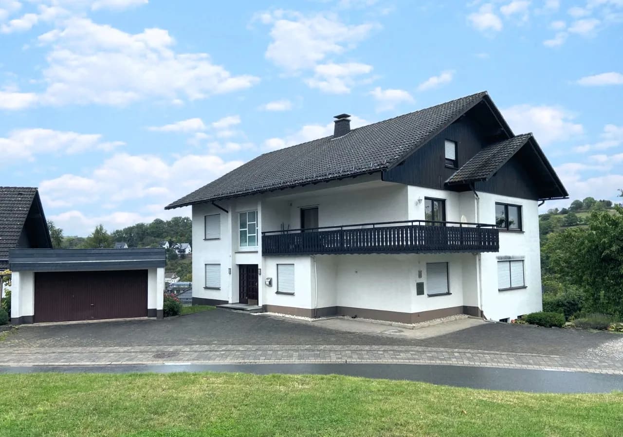Außenansicht des Wohnhauses mit weißer Putzfassade, dunkler Holzverkleidung im Giebelbereich, durchgehendem Obergeschossbalkon, Hauseingang, Garage und befestigtem Vorplatz.