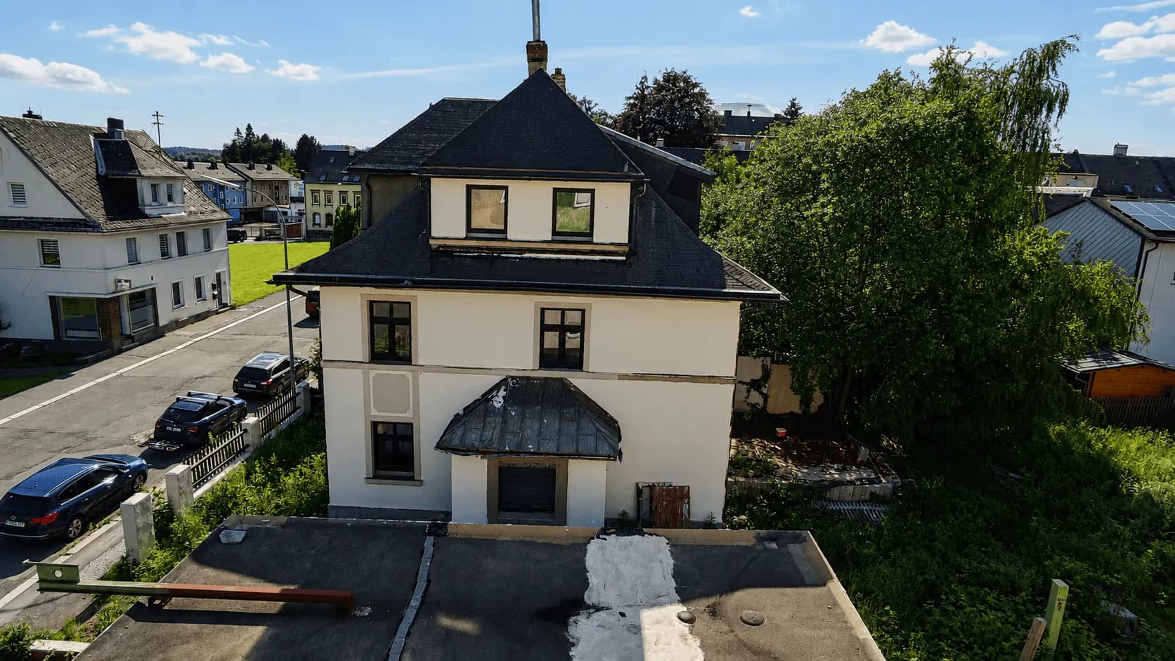 Erhöhte Außenansicht der Hausseite mit Mansarddach, Flachdach des Nebengebäudes/Garage und angrenzendem Gartenstück.