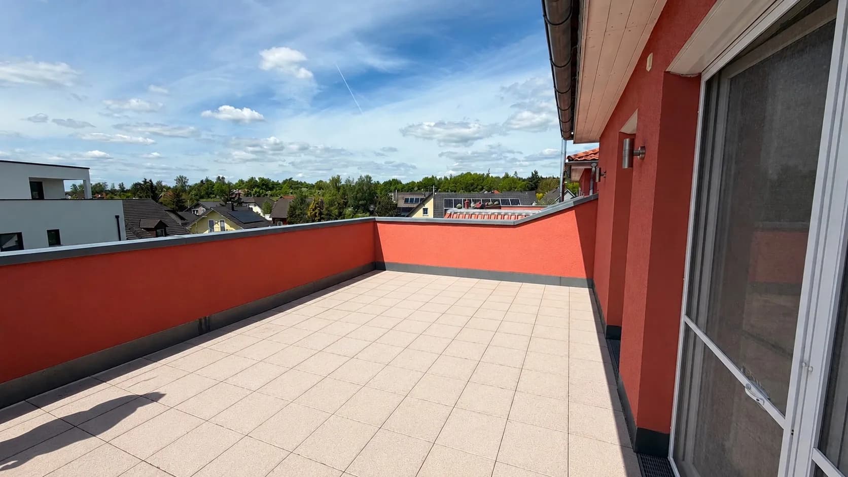 Weitläufige sonnige Dachterrasse im Obergeschoss mit hellem Plattenbelag, solider Brüstungsmauer und weitem Panoramablick über die Siedlung und Umgebung.
