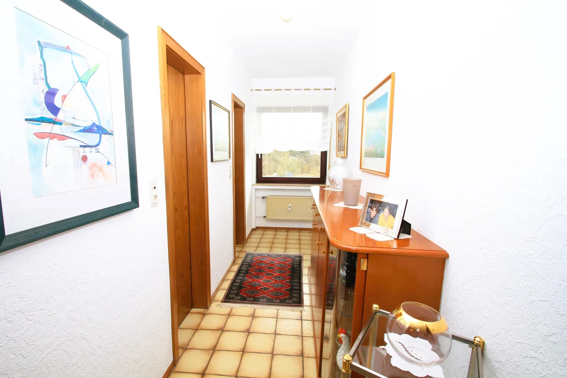Heller Hausflur mit weißem Wandputz, sandfarbenem Fliesenboden, klassischer Holzkommode samt Vitrine, gerahmten Bildern und Tageslichtfenster an der Stirnseite.