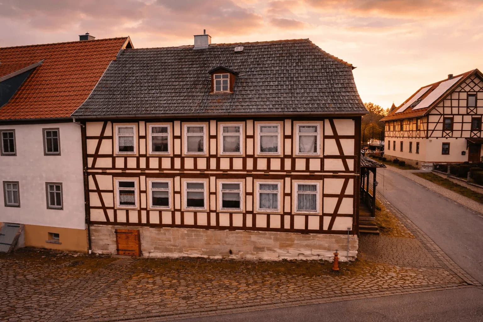 Atmosphärische Außenaufnahme des Fachwerkhauses im Abendlicht mit sichtbarem Holzfachwerk, Sandsteinsockel mit Kellerzugang und historischem Straßenpflaster.