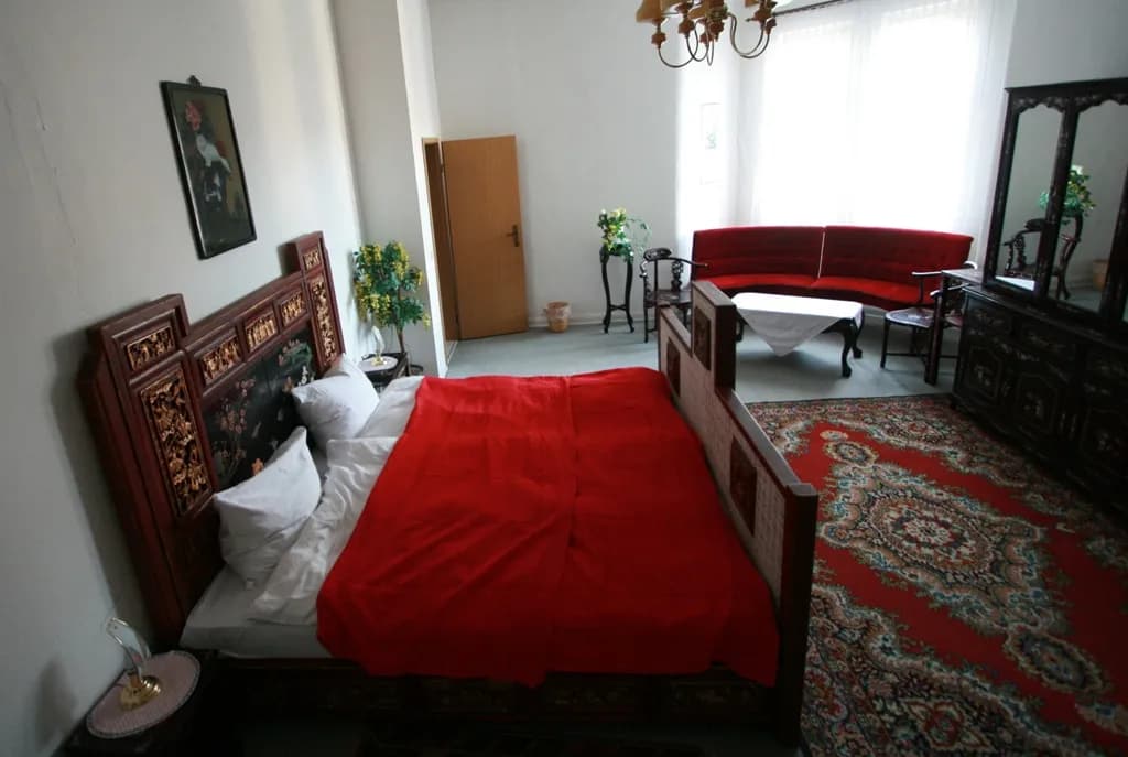 Großzügiges Themen-Schlafzimmer im asiatischen Stil mit kunstvoll verziertem Holzbett, rotem Orientteppich, gemütlicher Sitzecke mit Rundsofa und historischer Schminkkommode samt Spiegelelementen.