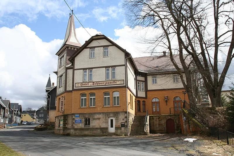 Straßenansicht des historischen Hotelgebäudes „Schwarzaquelle“ in Scheibe-Alsbach mit markantem Ecktorturm, Natursteinsockel, Zierverschieferung und repräsentativer Außentreppe.