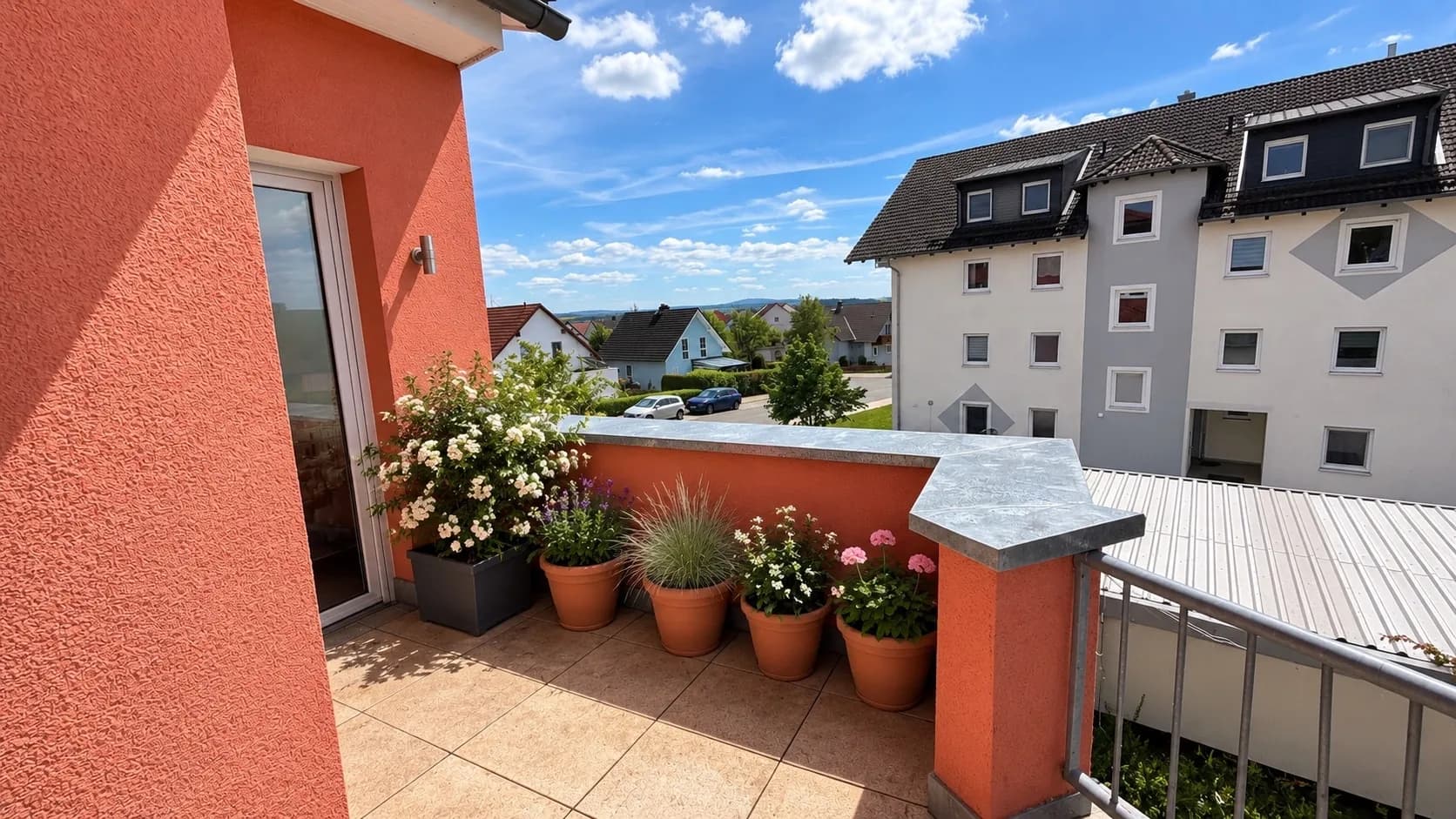 Gemütlicher Terrassenbereich mit Fliesenboden, blühenden Pflanzkübeln, robustem Metallgeländer und Ausblick auf das Wohnviertel.