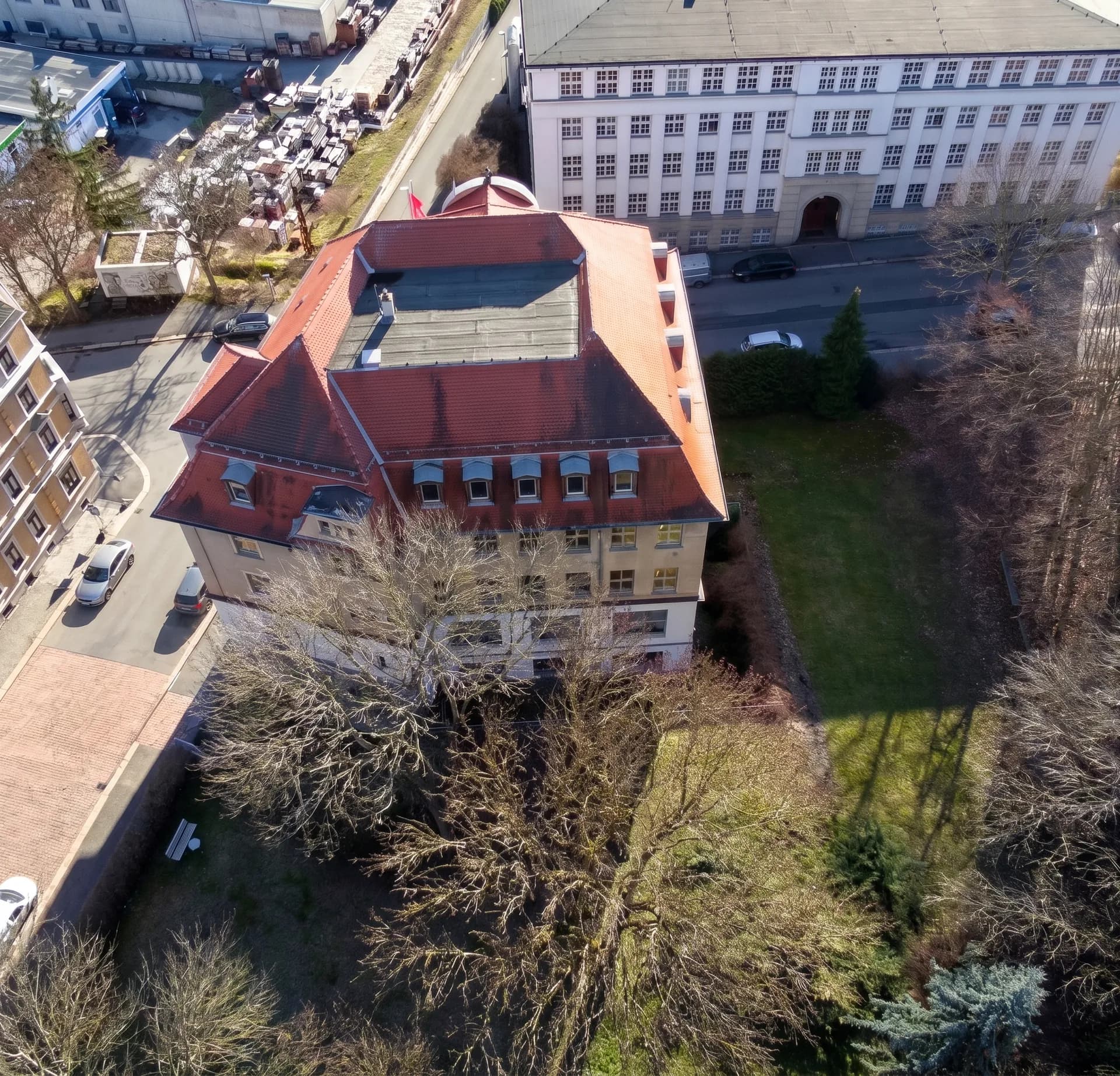 Luftaufnahme des repräsentativen, mehrgeschossigen Gebäudekomplexes mit markantem Ziegelmansarddach, Gauben und angrenzender Grünanlage.