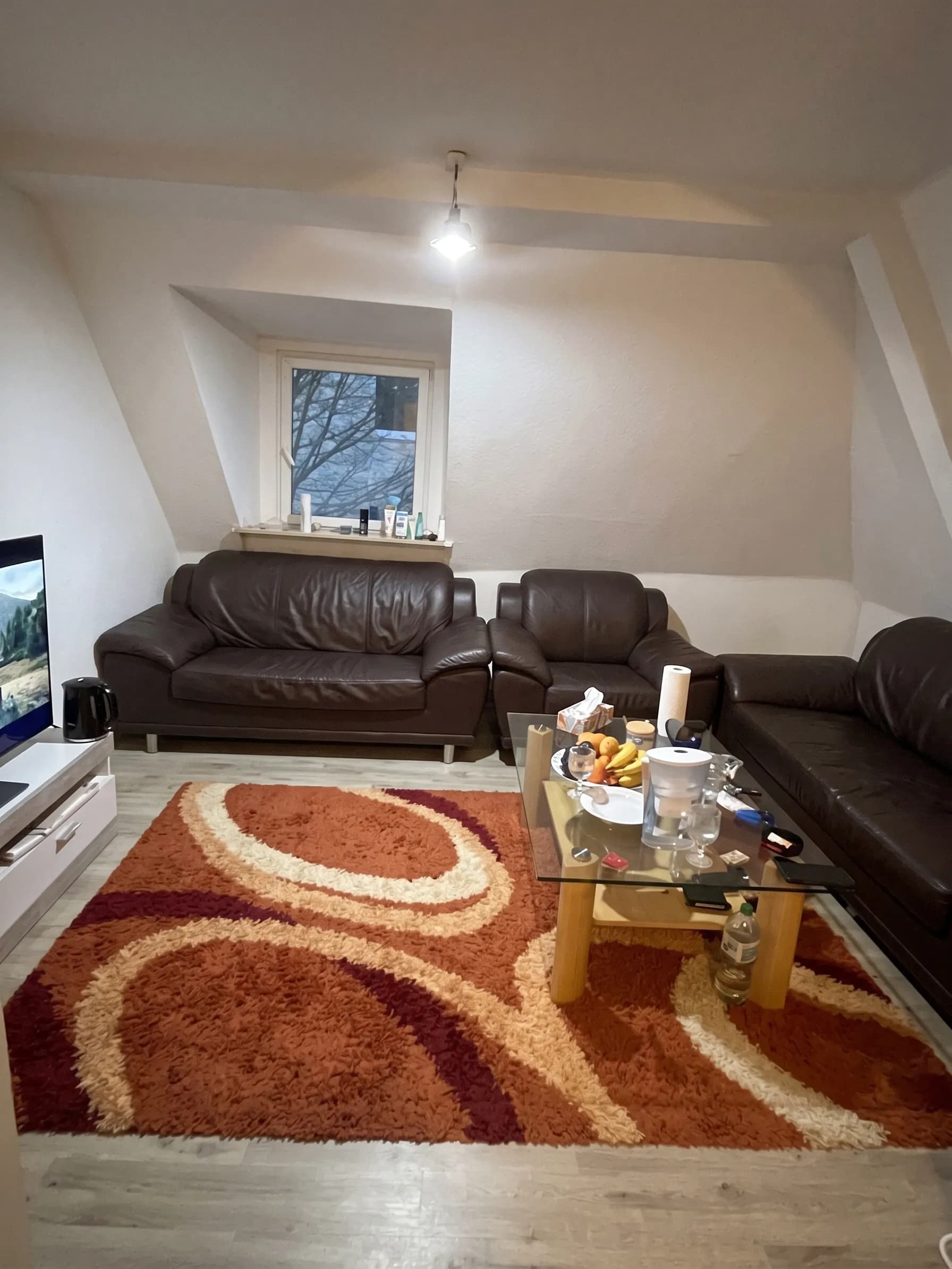Gemütliches Wohnzimmer im Dachgeschoss mit dunkler Leder-Polstergarnitur, Glas-Couchtisch, TV-Board und farbenfrohem Hochflorteppich.