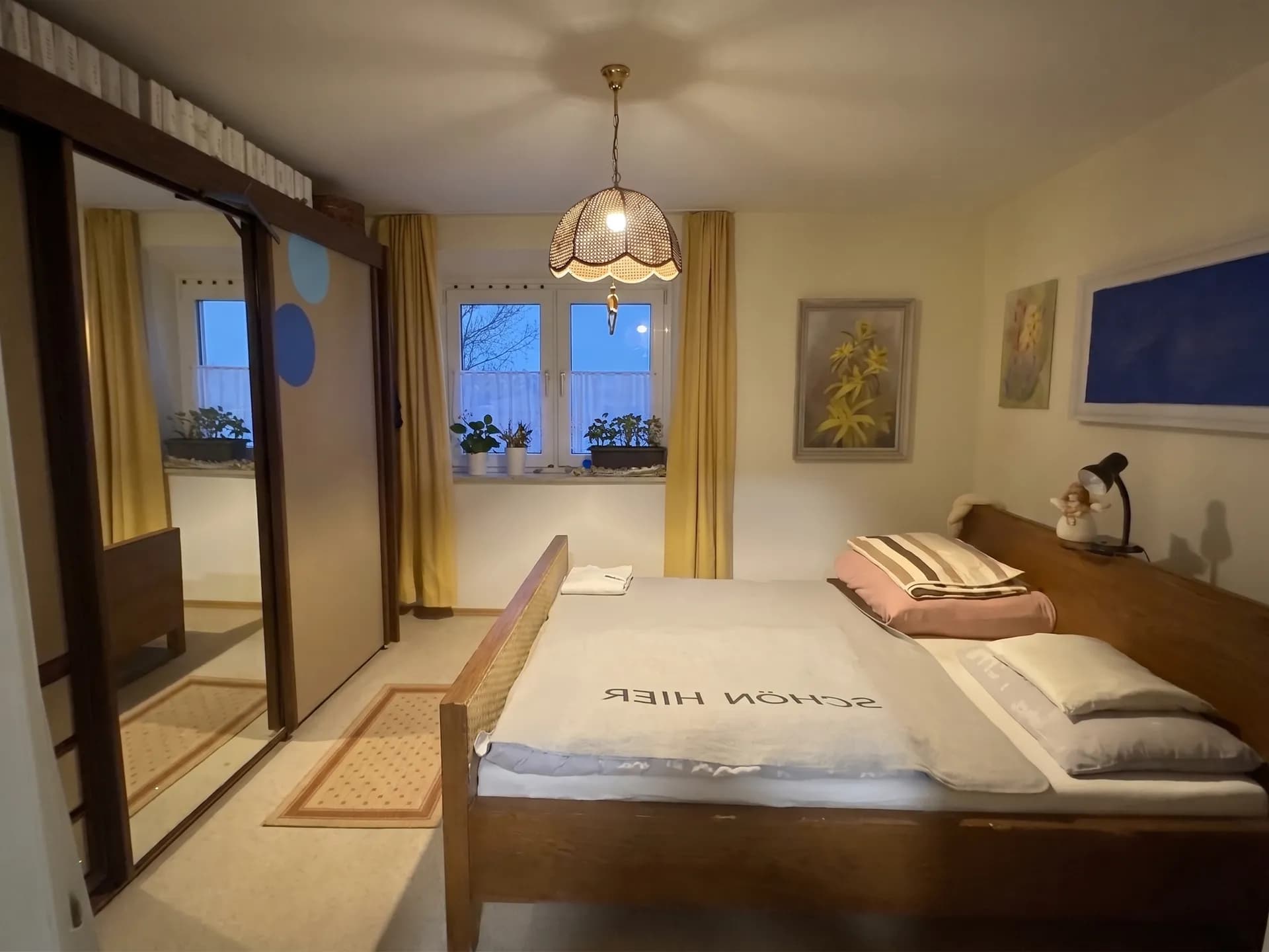 Gemütliches Schlafzimmer mit massivem Holzbett, großem Schiebetürenschrank mit Spiegelfronten und Tageslichtfenster mit wohnlichen gelben Vorhängen.