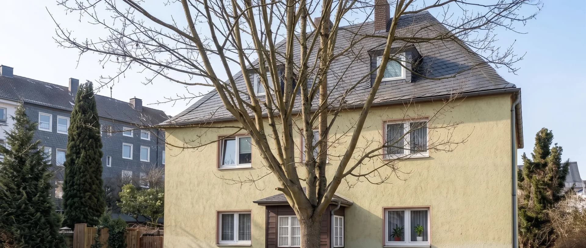 Außenansicht des gepflegten Mehrfamilienhauses mit ausgebautem Dachgeschoss, holzverkleidetem Eingangsbereich, Vorplatz und altem Baumbestand.
