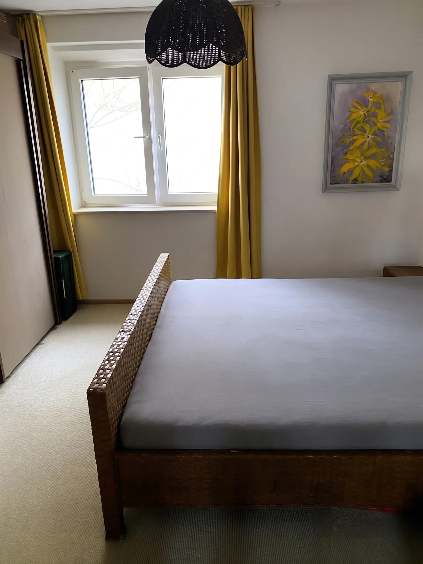 Gemütliches Schlafzimmer mit Geflecht-Doppelbett, Schiebetürenschrank, Wandbild und Tageslichtfenster mit gelben Vorhängen.