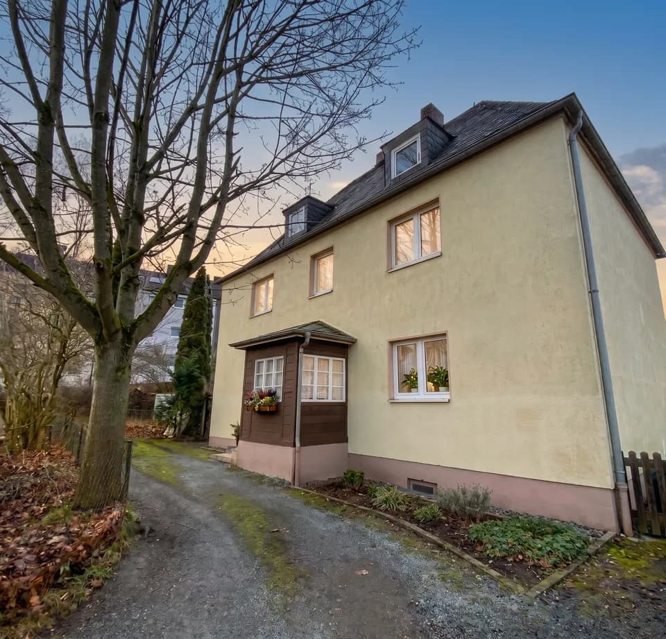 Perspektivische Außenansicht des Mehrfamilienhauses mit Zufahrt, holzverkleidetem Windfang, Dachgauben und natürlichem Baumbestand.
