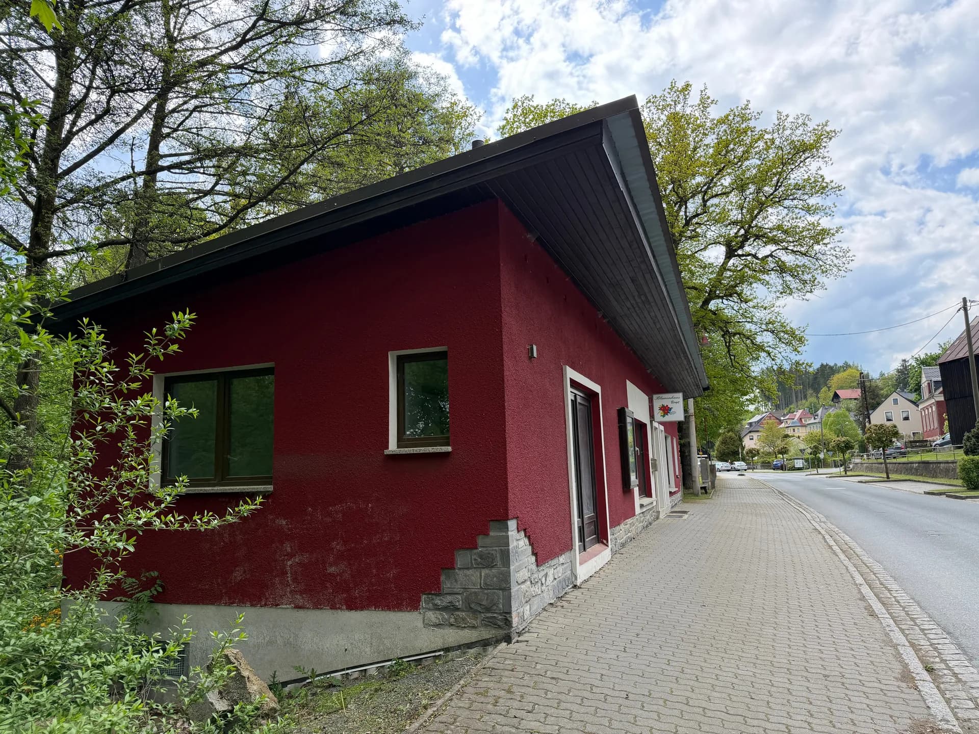 Perspektivische Außenansicht des Gebäudes entlang des gepflasterten Gehwegs mit seitlichen Fenstern und herrlich grünem Baumbestand.