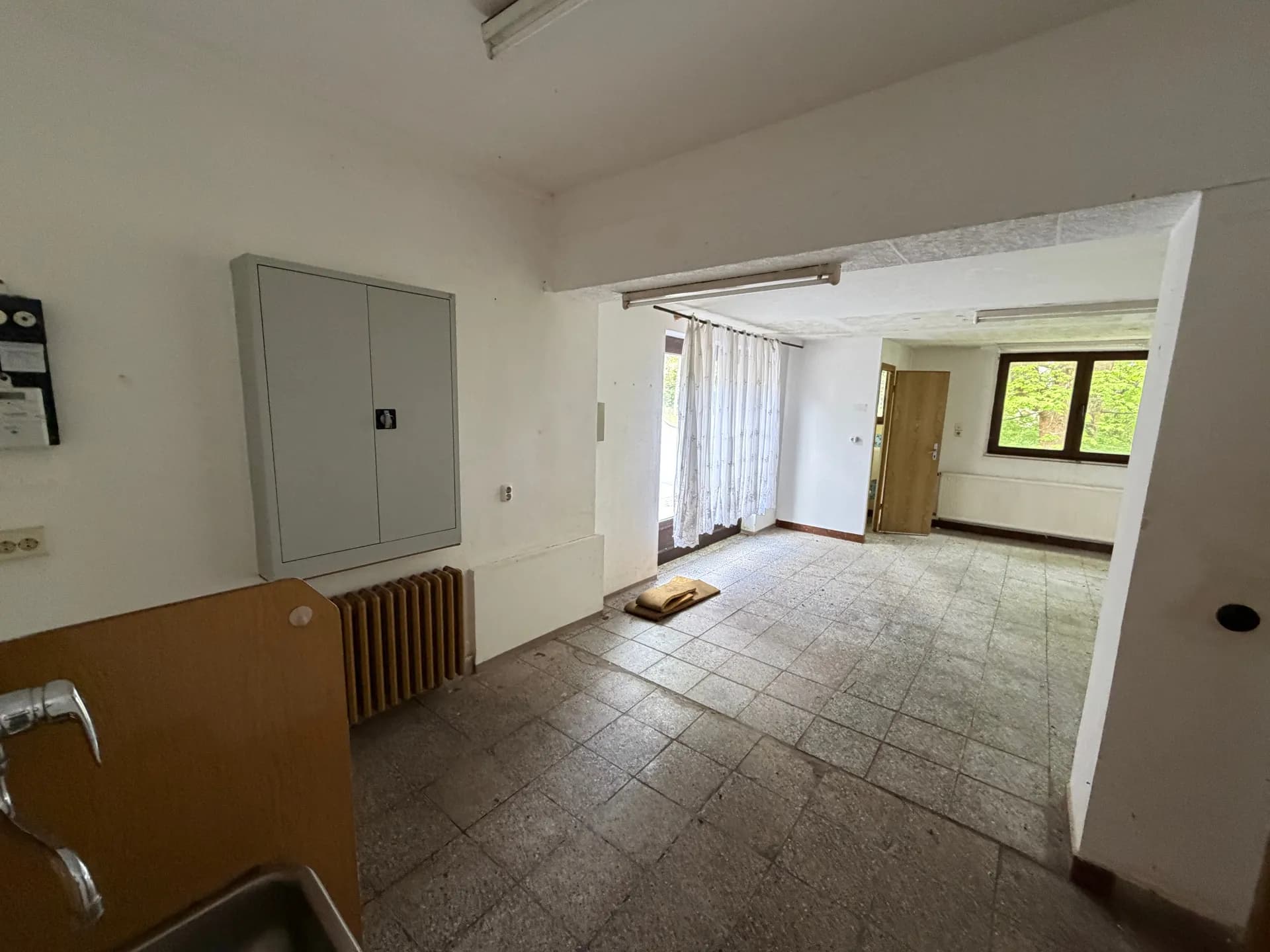 Perspektivische Außenansicht des Gebäudes entlang des gepflasterten Gehwegs mit seitlichen Fenstern und herrlich grünem Baumbestand.