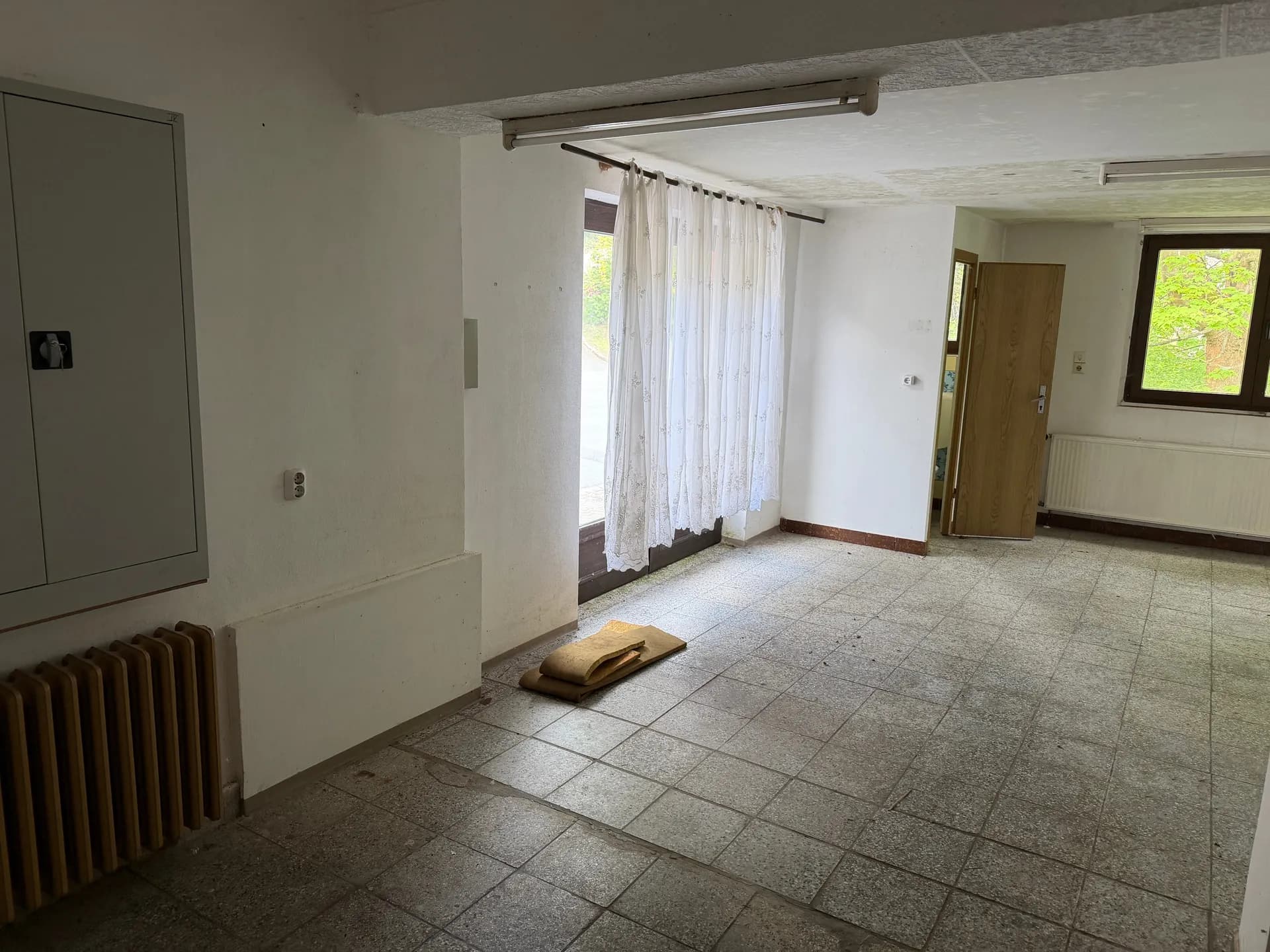 Großzügiger, heller Raum mit Terrazzo-Fliesenboden, großem Fenster mit Blick ins Grüne, Heizkörper und Zugang zu weiteren Räumlichkeiten.