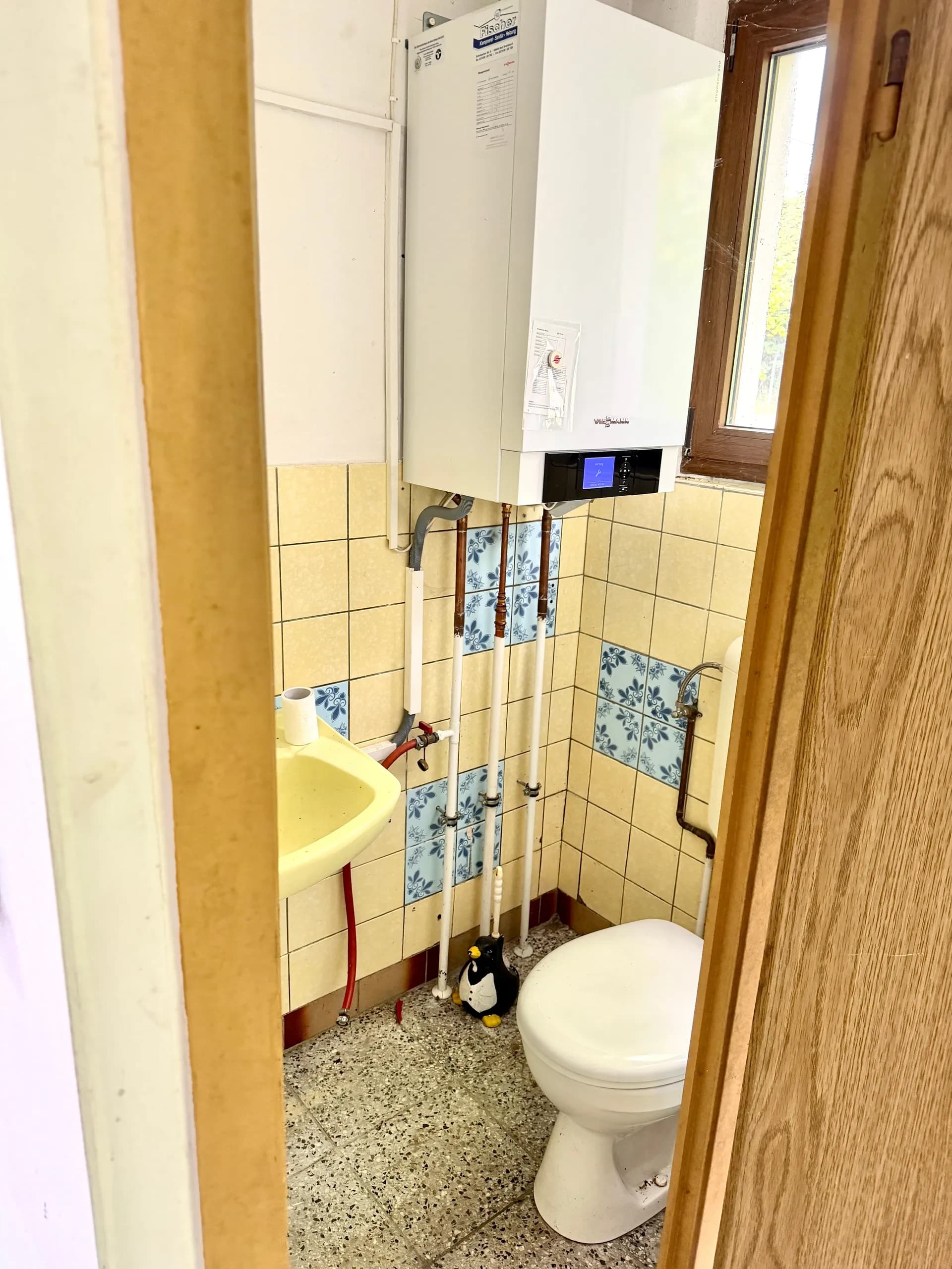 Kompaktes WC / Technikraum mit wandhängender Fischer-Gastherme, Handwaschbecken, WC, Wandfliesen und Lüftungsfenster.