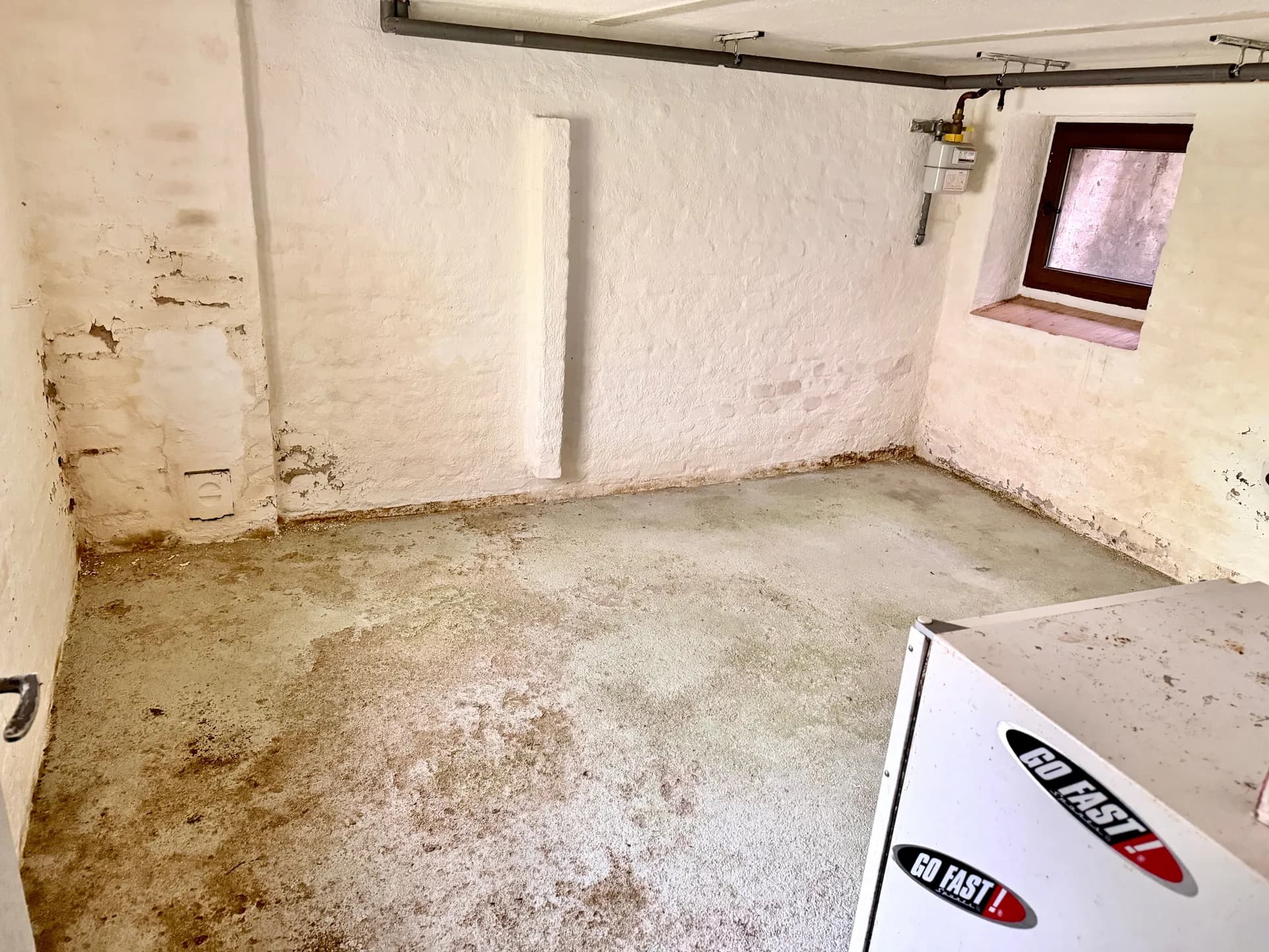 Trockener Kellerraum mit weiß gestrichenem Mauerwerk, Betonboden, Tageslicht-Kellerfenster und anliegenden Versorgungsleitungen.