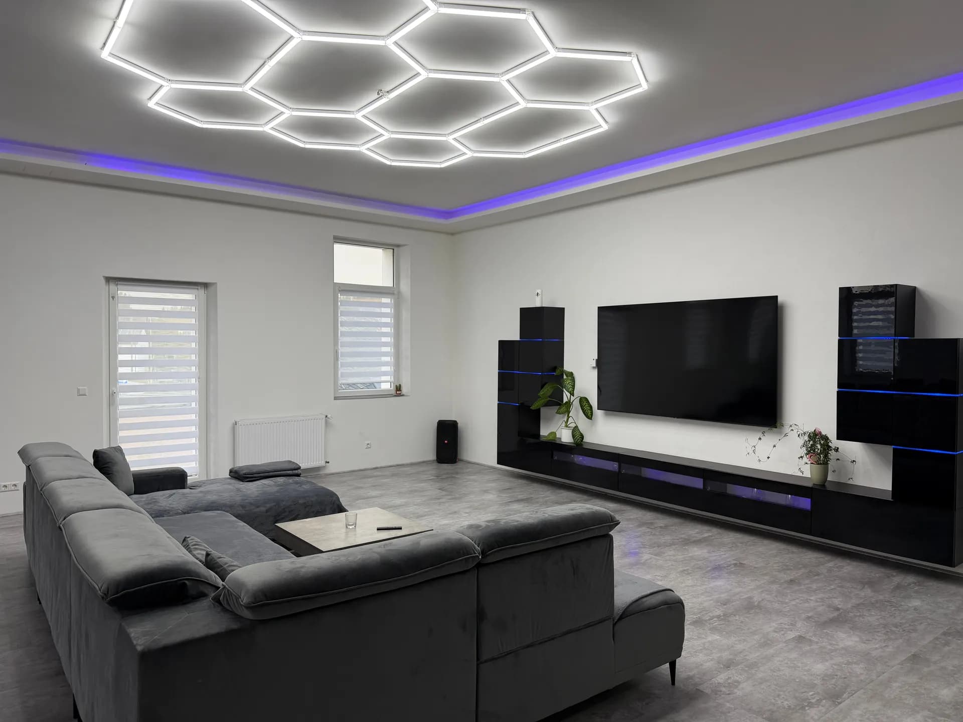 Moderner Wohnbereich mit ausladender Eckcouch, hinterleuchteter schwarzer TV-Wand, großem Flachbildfernseher und Waben-LED-Deckenbeleuchtung.