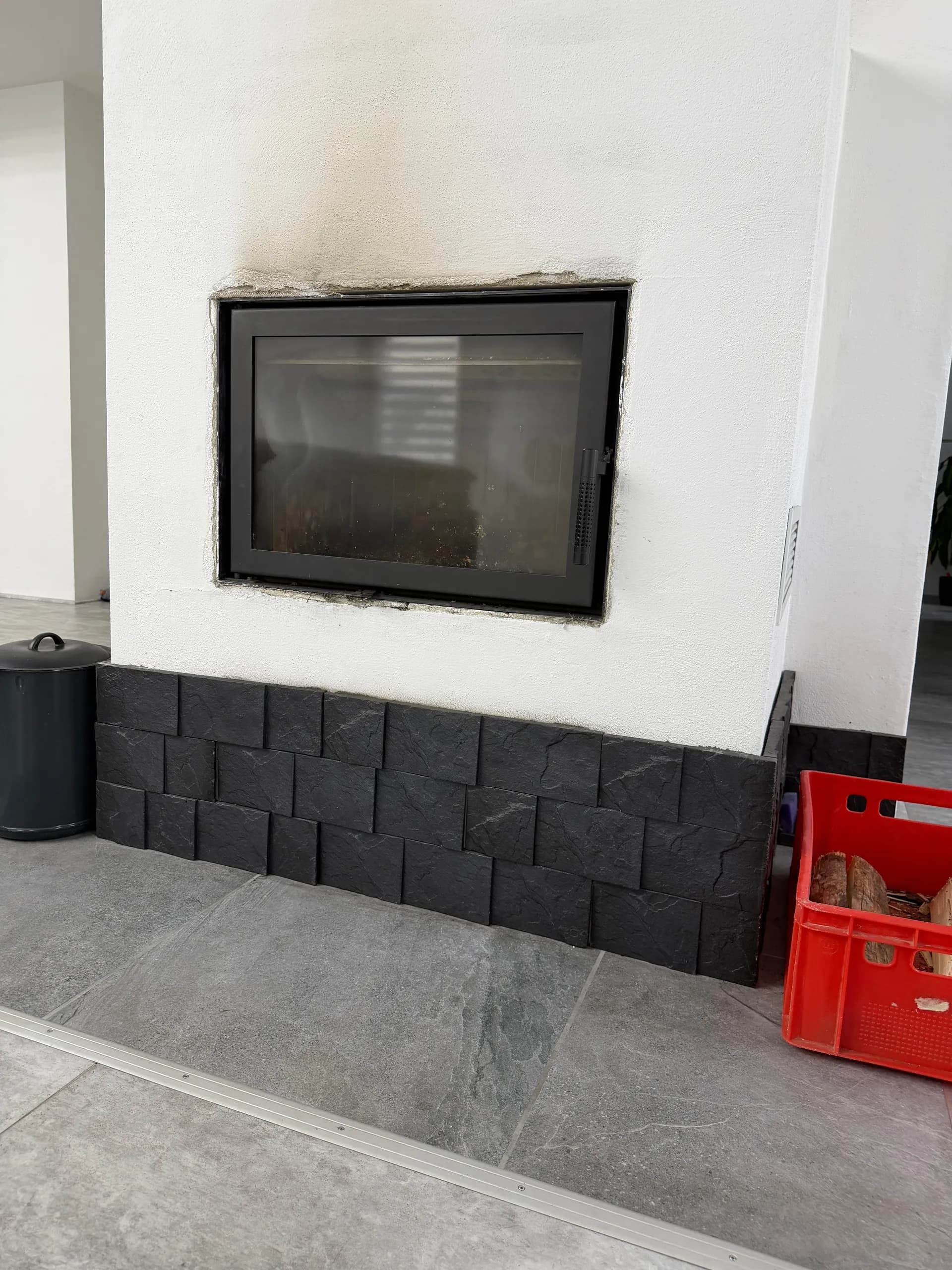Moderner geschlossener Kamin mit Sichtscheibe, dunkler Naturstein-Sockelverkleidung und Kaminholzablage.