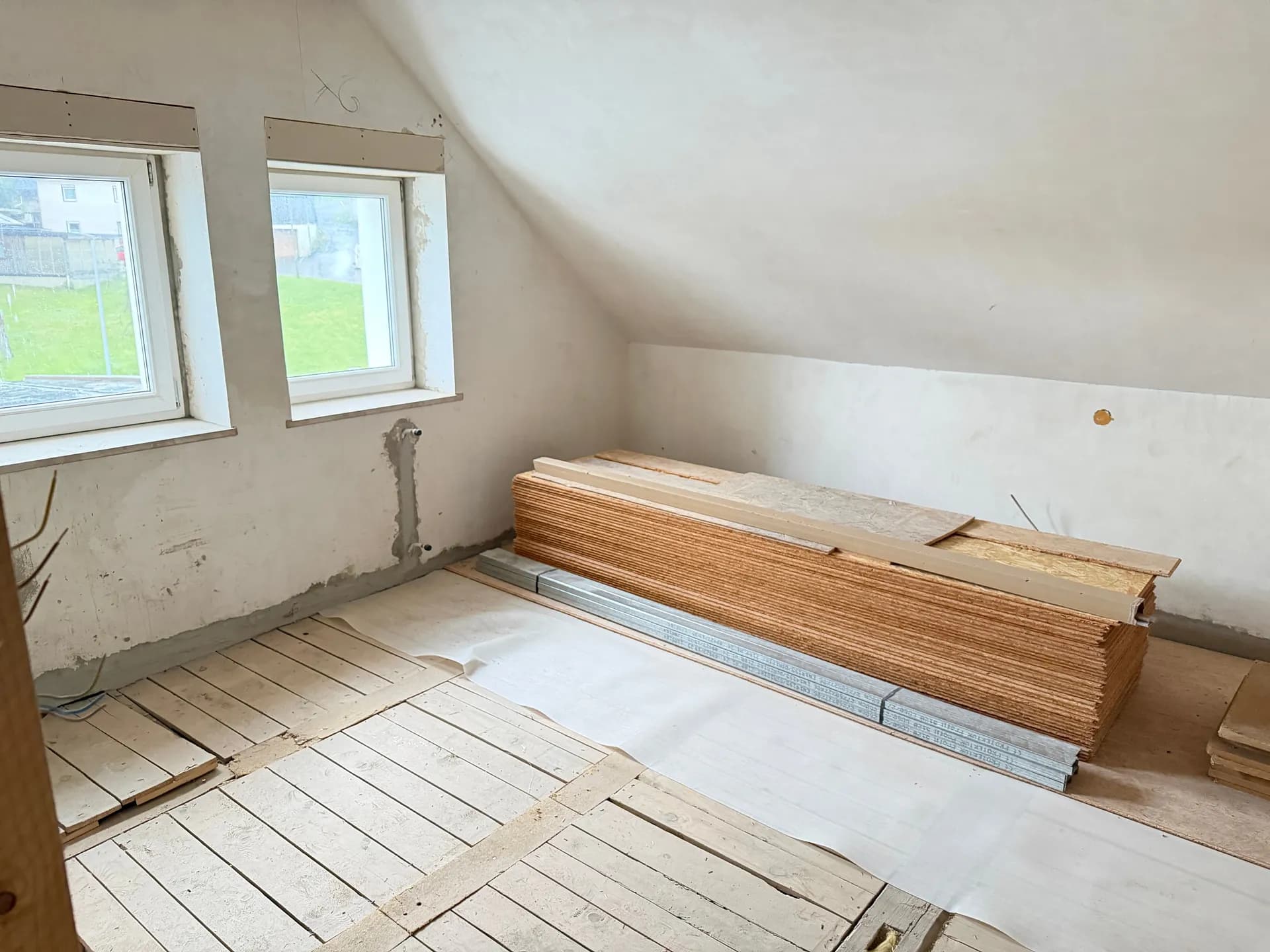 Ausbaufähiges Dachgeschosszimmer mit Dachschrägen, zwei Fenstern und gelagerten Baumaterialien zur individuellen Fertigstellung.