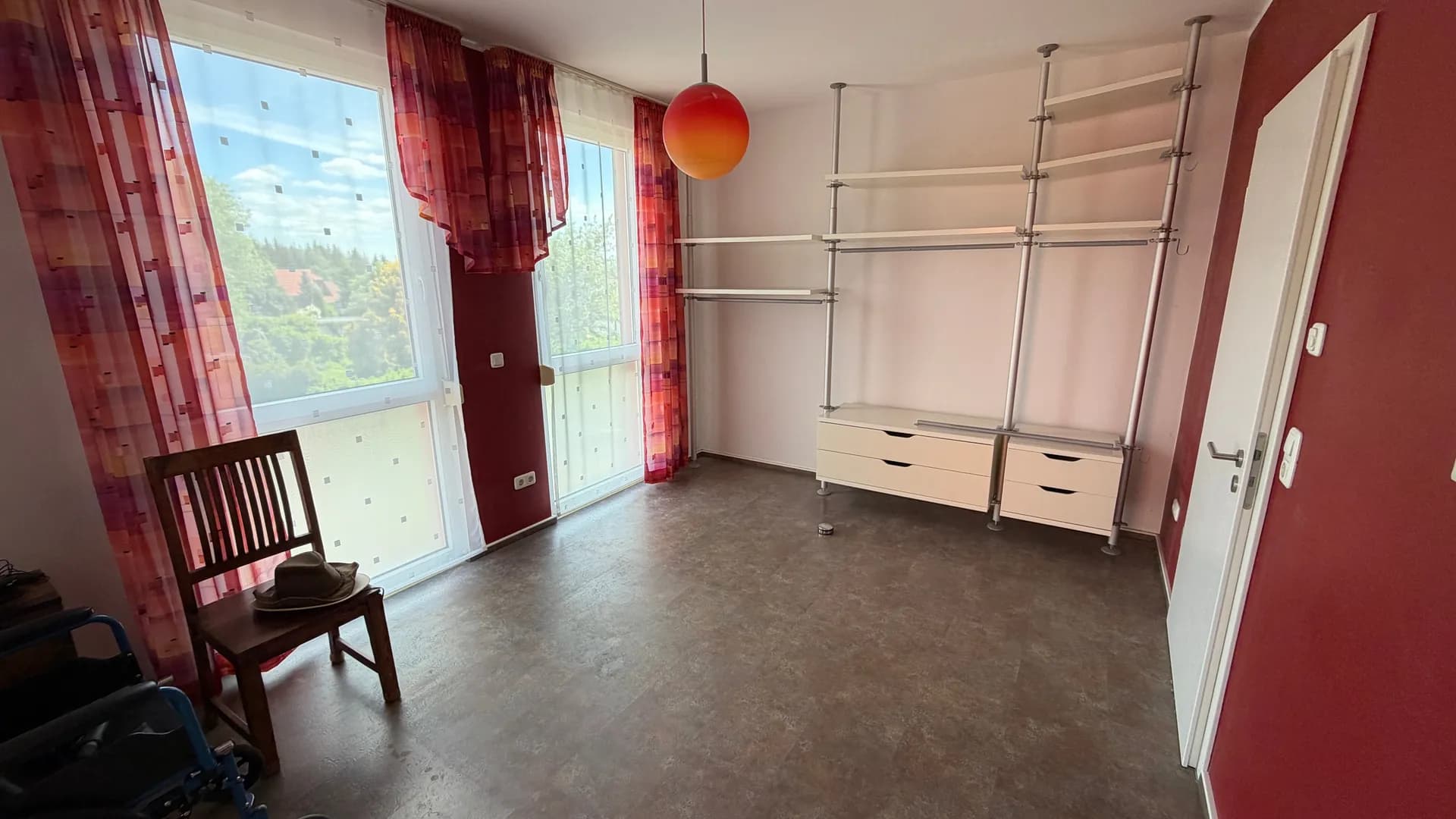 Lichtdurchflutetes Zimmer mit bodentiefen Fenstern, modernem offenen Regalsystem mit Kommodenauszügen und weinroter Akzentwand.