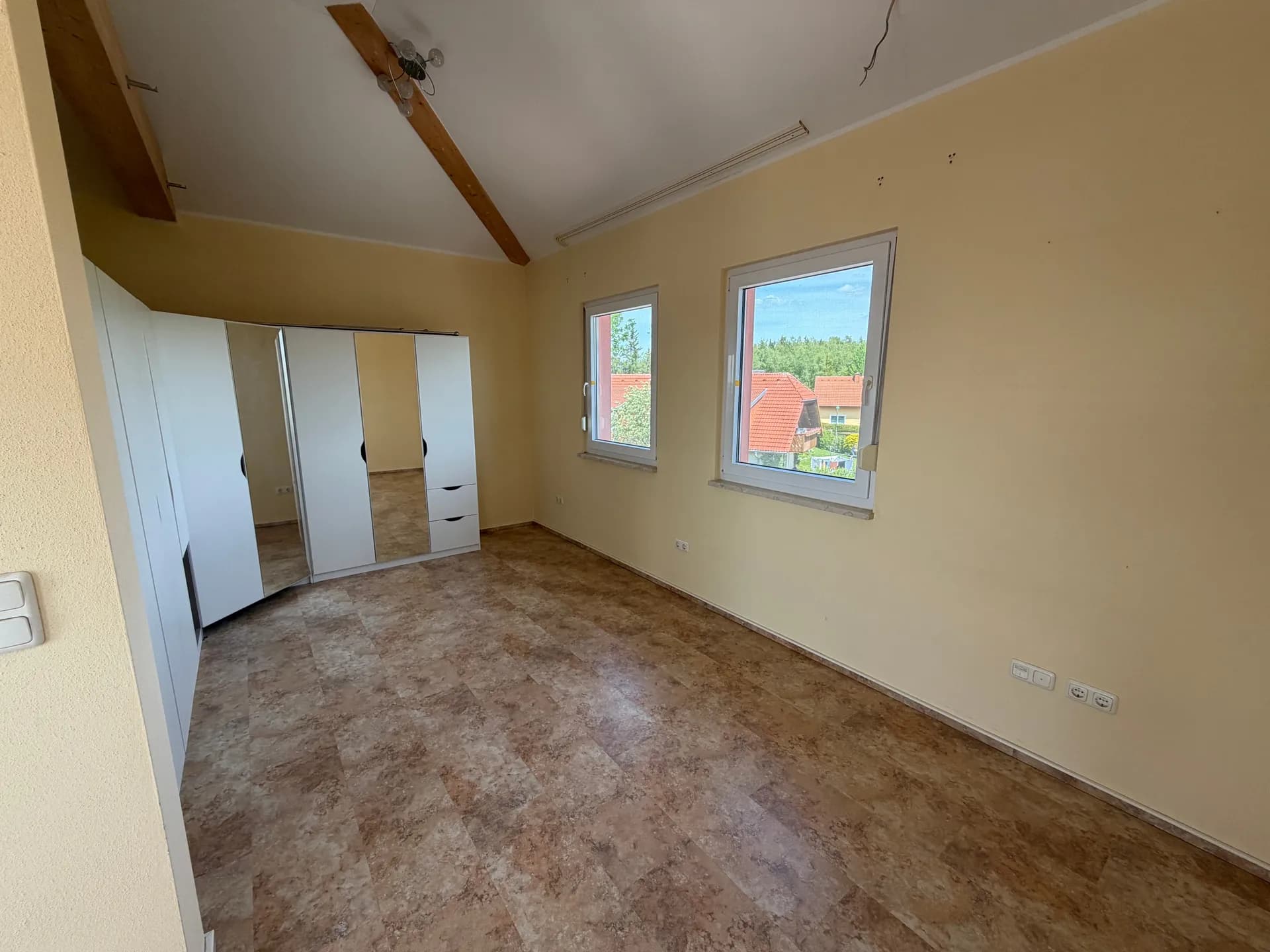 Großes, lichtdurchflutetes Schlafzimmer mit hoher Decke, sichtbarem Holzbalken, zwei Fenstern, Bodenbelag in Steinoptik und geräumigem Spiegelschrank.