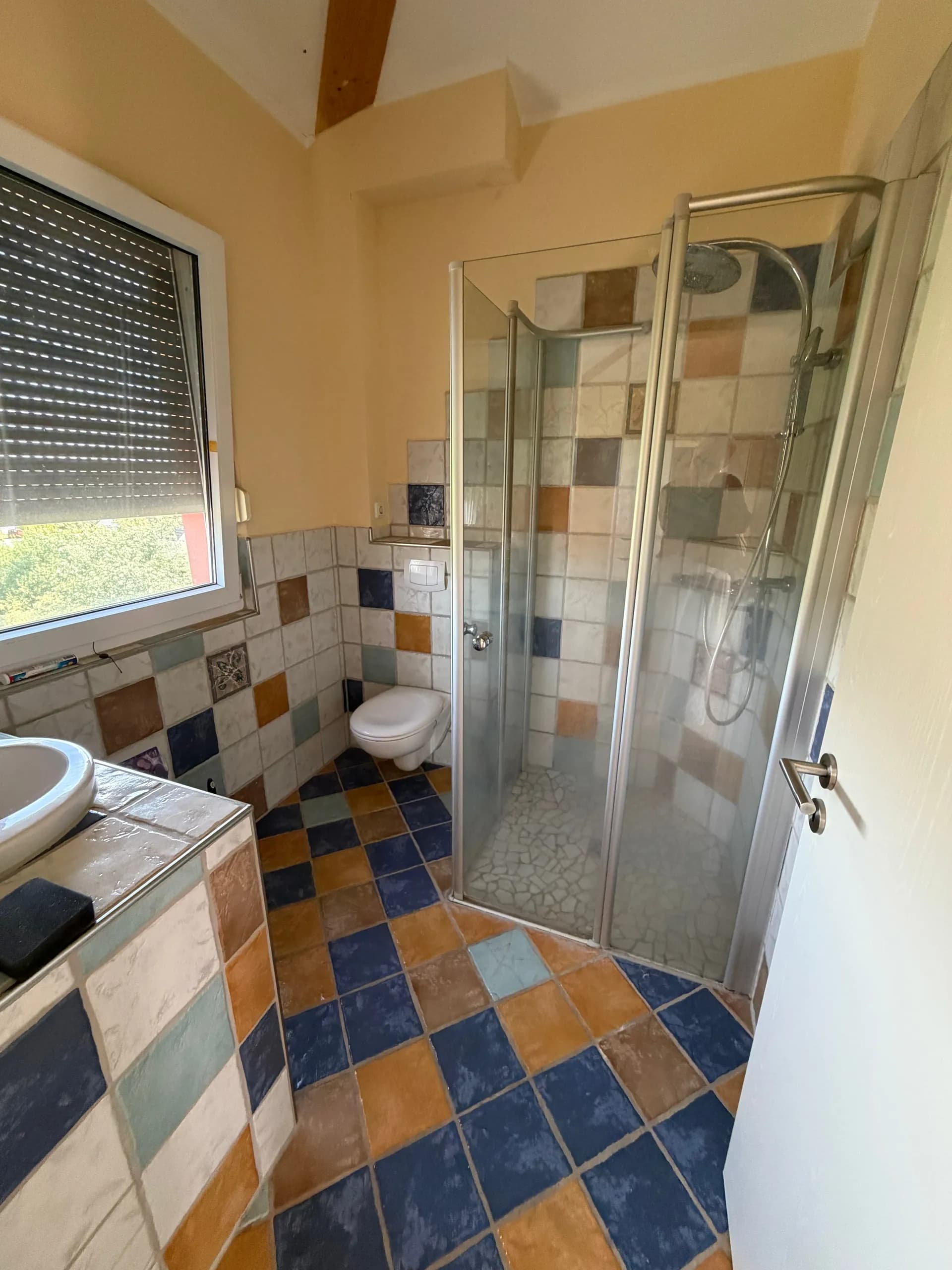 Badezimmer im mediterranen Stil mit bunten Mosaikfliesen, Glas-Duschkabine, Hänge-WC, Waschtisch und Fenster inklusive Rollladen.