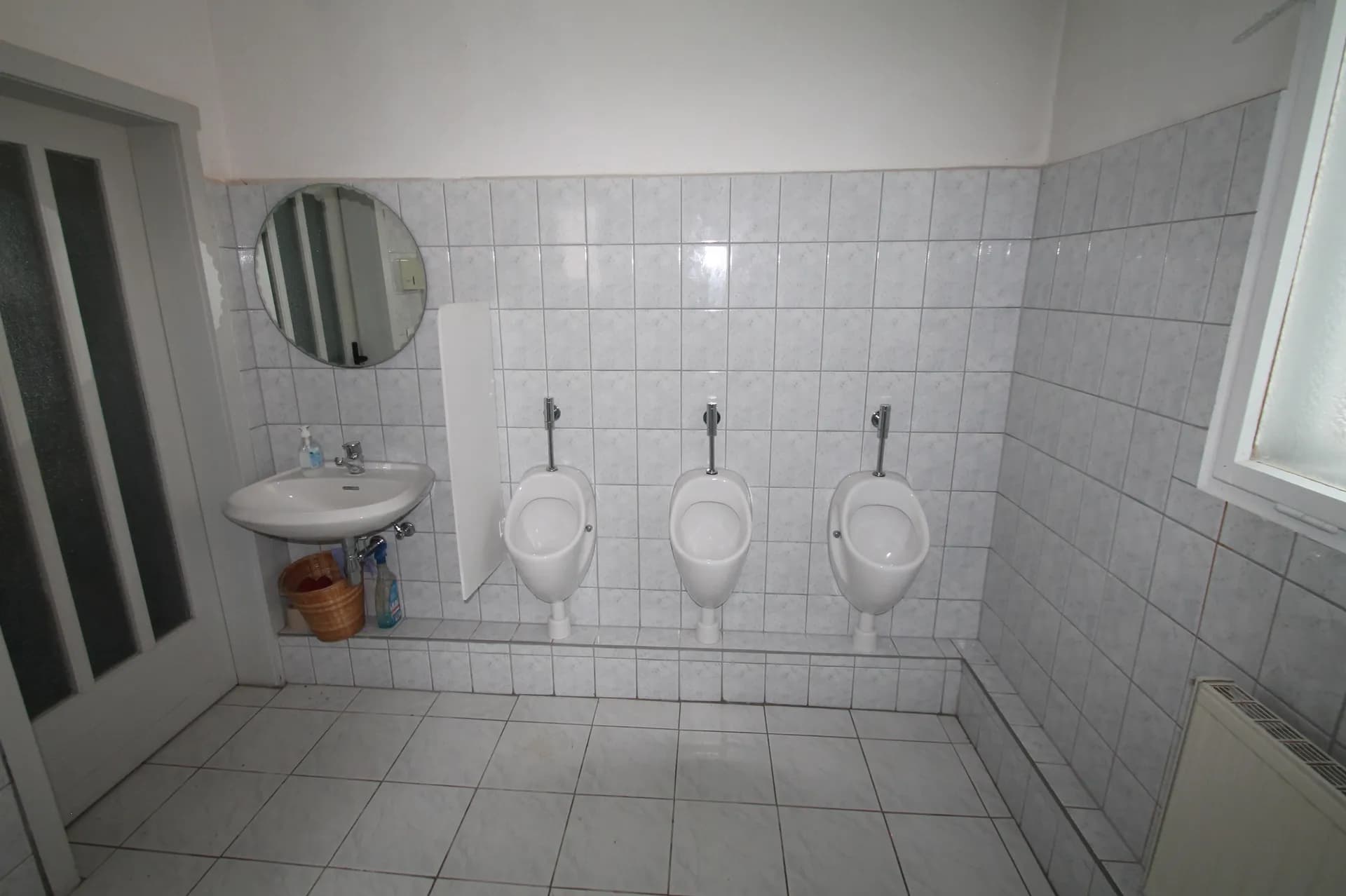 Herren-WC-Anlage mit Fliesenbelag, drei Wand-Urinalen samt Trennwand, Handwaschbecken, Spiegel und Heizkörper.