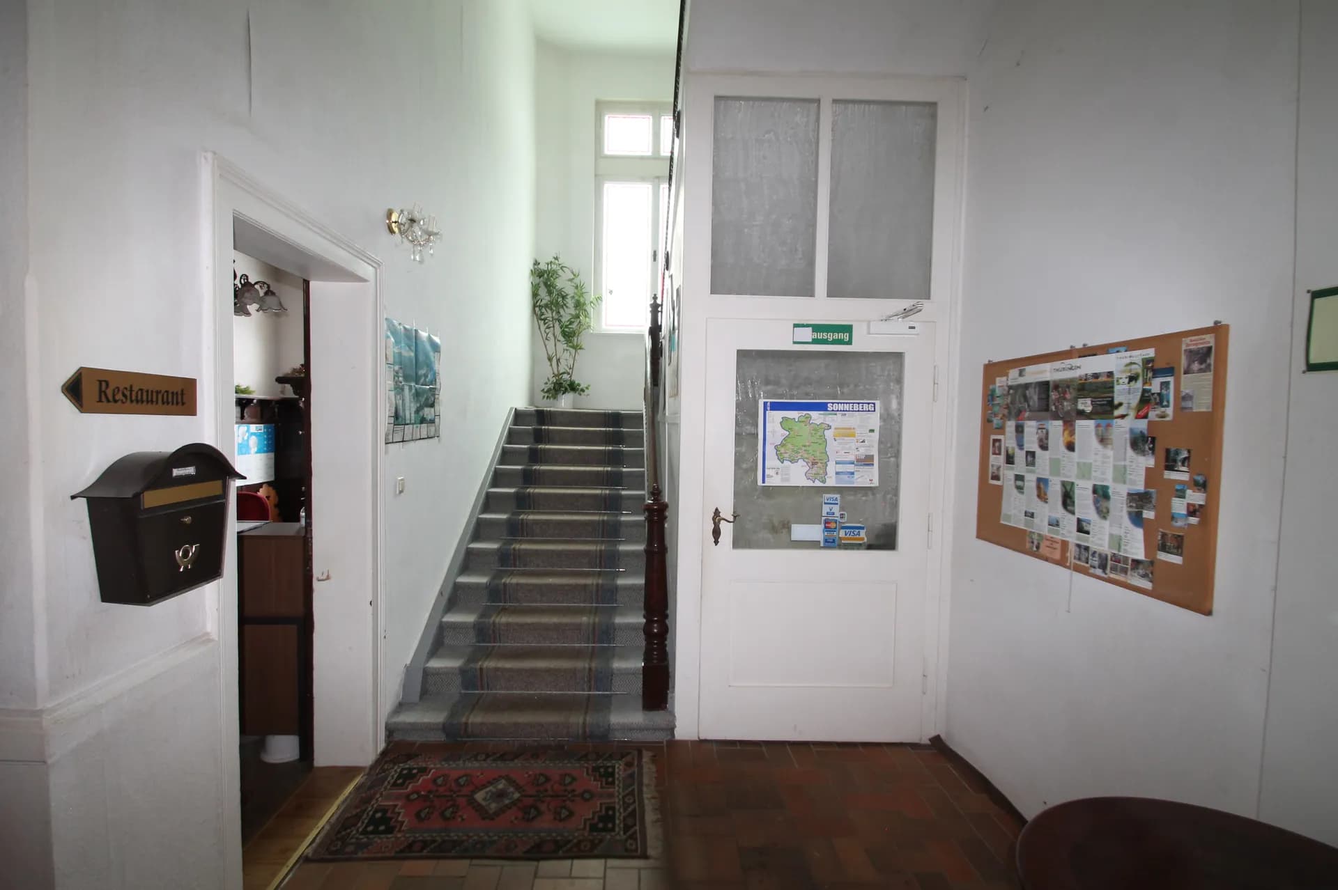 Eingangsfoyer des Hotels mit historischem Treppenaufgang samt Läufer, Informationstafel, Ausgangstür und Wegweiser zum Restaurantbereich.