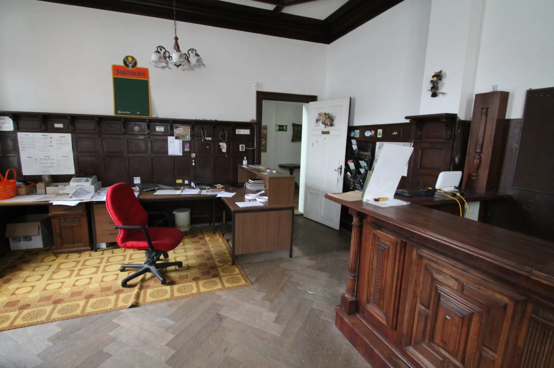 Klassischer Hotel-Rezeptionsbereich mit massiver Holztheke, Wandvertäfelung, Schreibtisch-Arbeitsplatz samt rotem Bürostuhl und Fischgrätparkett.