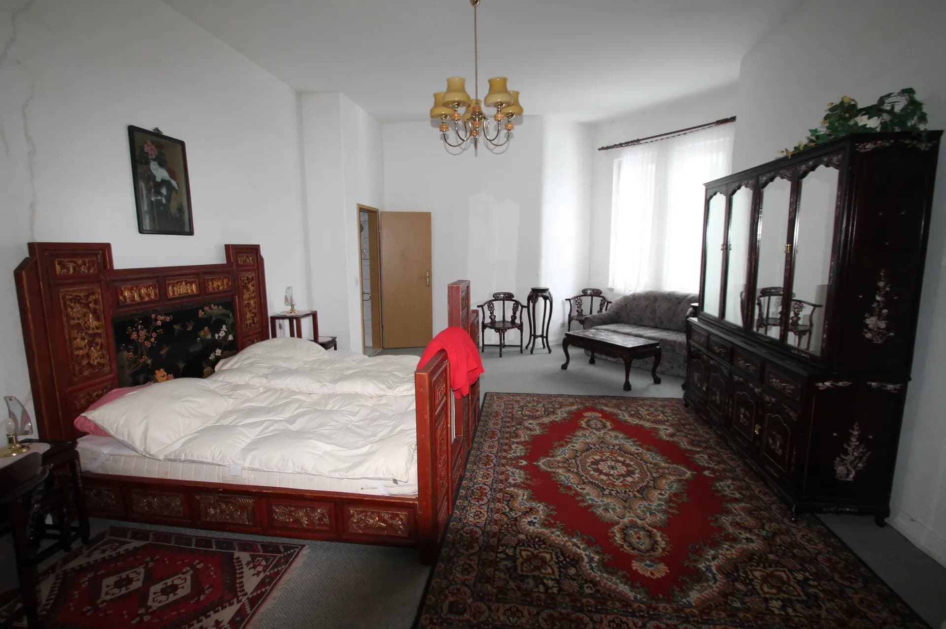 Geräumiges Themen-Schlafzimmer im asiatischen Stil mit kunstvoll verziertem Holz-Doppelbett, großem Orientteppich, Sitzecke mit Polstersofa und antikem Spiegelschrank.