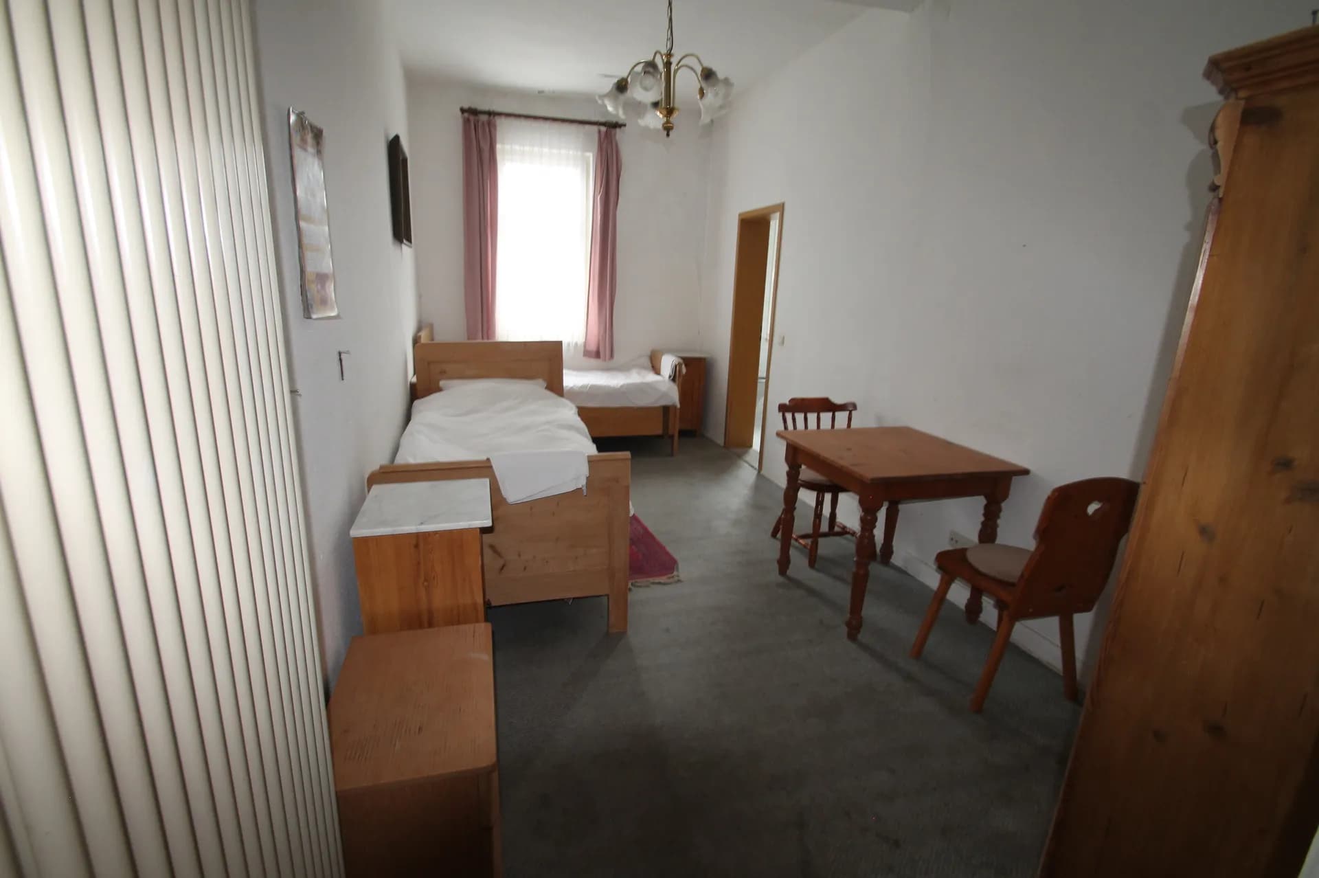 Helles Hotel-Zweibettzimmer mit zwei Einzel-Holzbetten, Nachttischen, schlichtem Holztisch samt Stühlen und hohem Fenster mit Vorhängen.