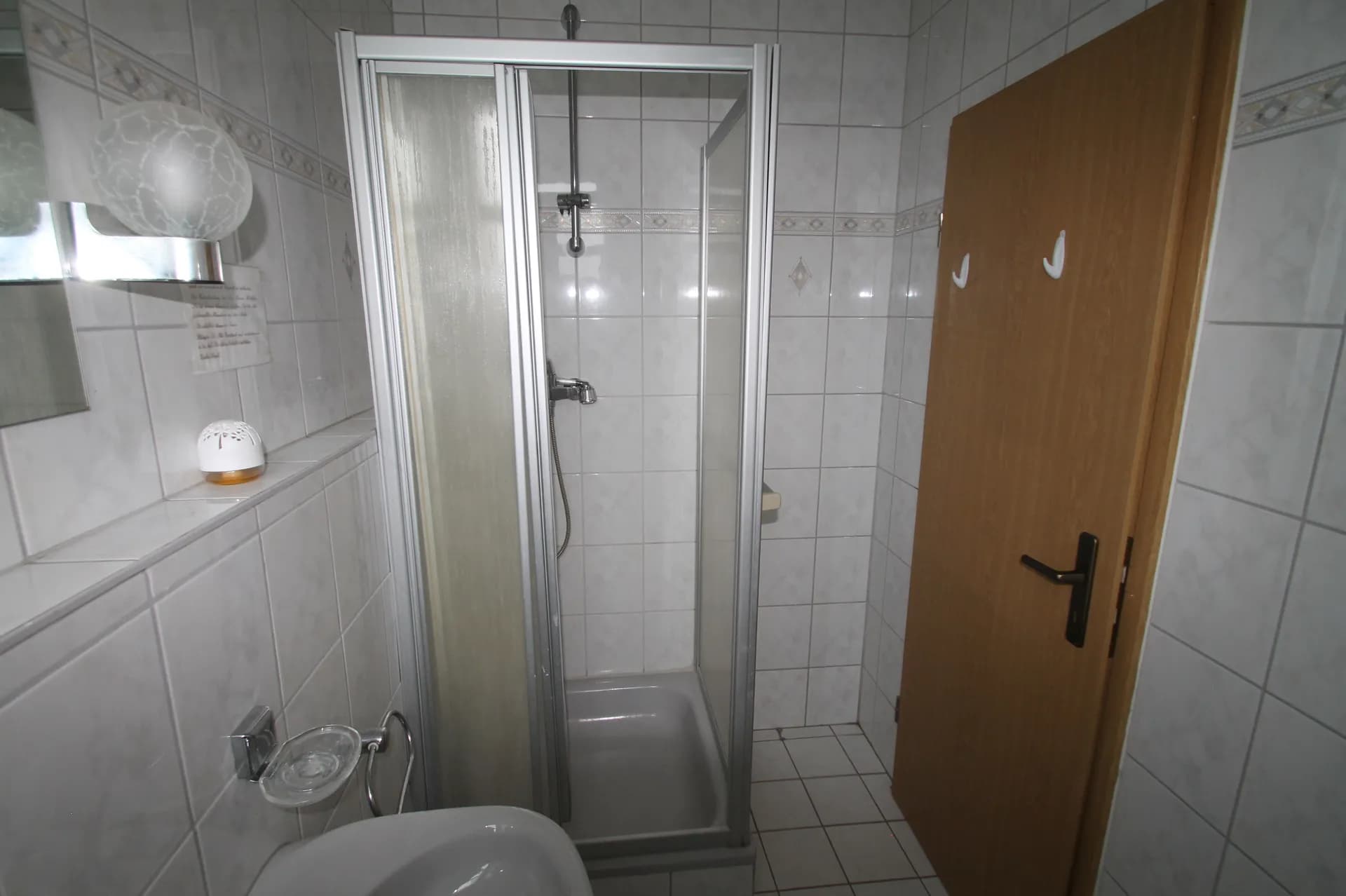 Kompaktes Hotelbad mit weißer Wandverfliesung, Eckduschkabine samt Schiebetüren, Waschbecken, Kugel-Wandleuchte und Holztür.
