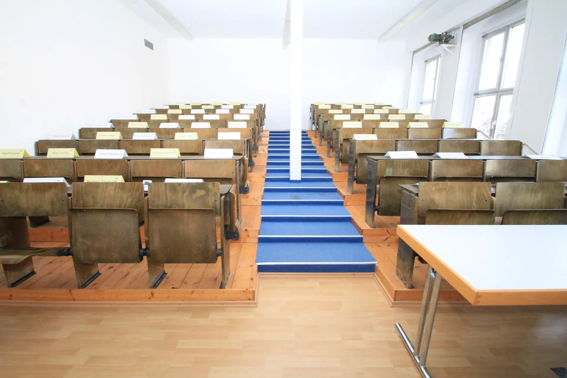 Klassischer Hörsaal mit ansteigender Holzbestuhlung, blau ausgelegtem Mittelgang, Dozententisch und großen Tageslichtfenstern.