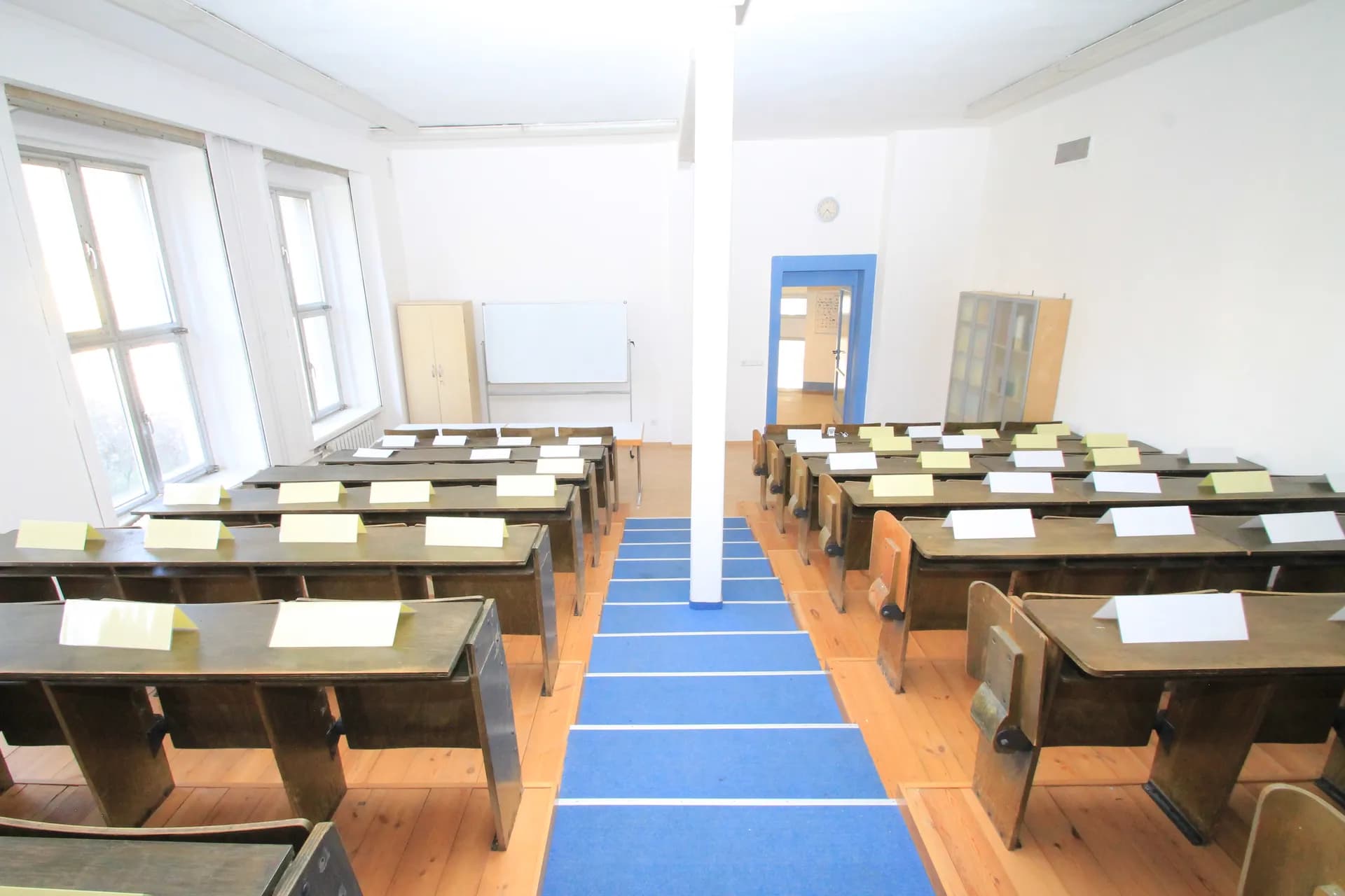 Klassischer Hörsaal mit ansteigenden Holzbankreihen, blauem Teppichläufer im Mittelgang, Whiteboard im Dozentenbereich und hohen Fenstern.