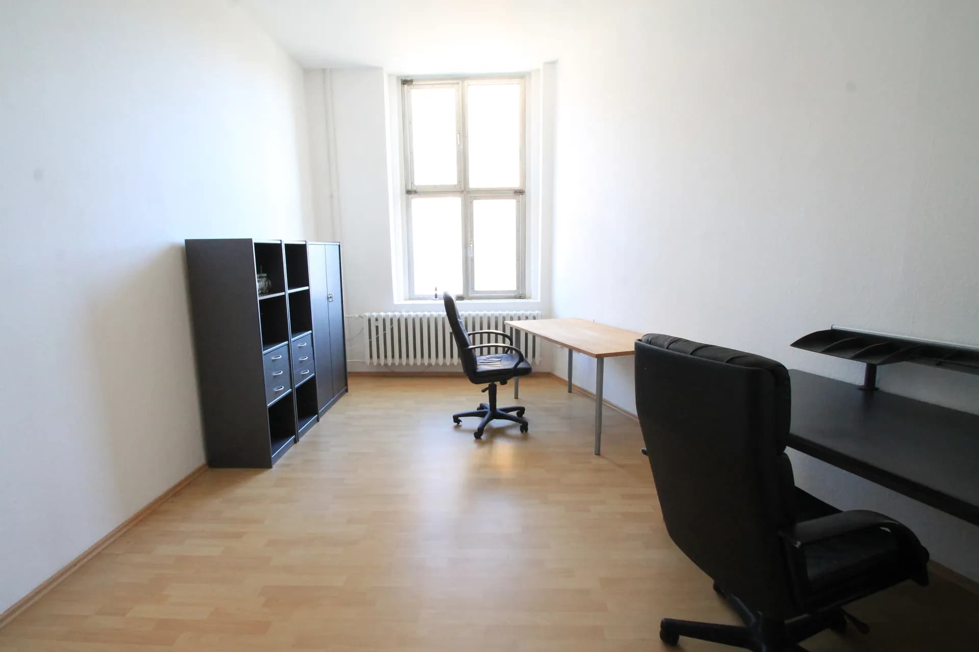 Helles Einzel- oder Doppelbüro mit Laminatboden, Schreibtischen, Drehstühlen, Aktenschrank und hohem Fensterelement.