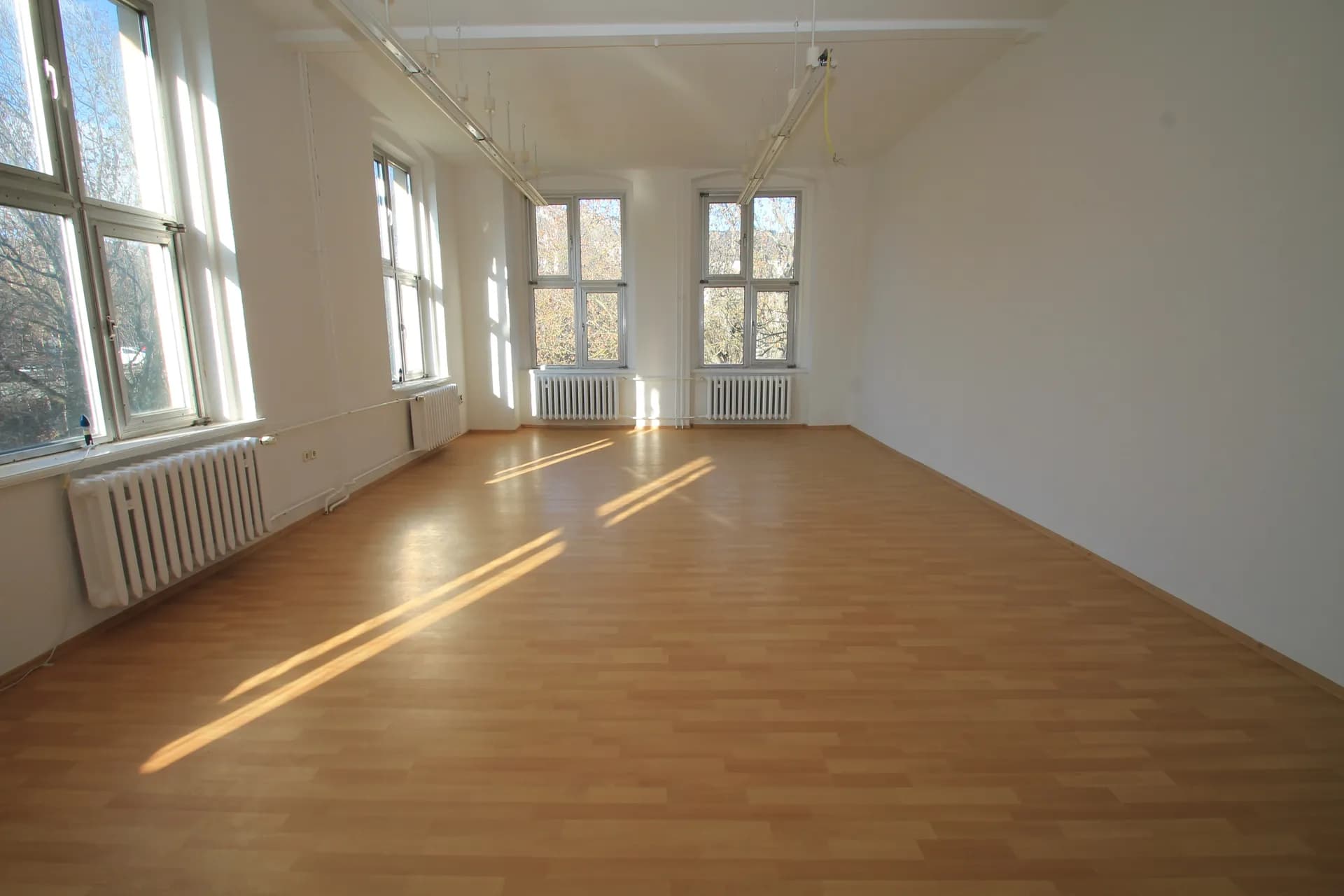 Großzügiger Schulungs- oder Büroraum mit warmem Laminatboden, Deckenbeleuchtung und vier hohen Fensterelementen für optimalen Lichteinfall.