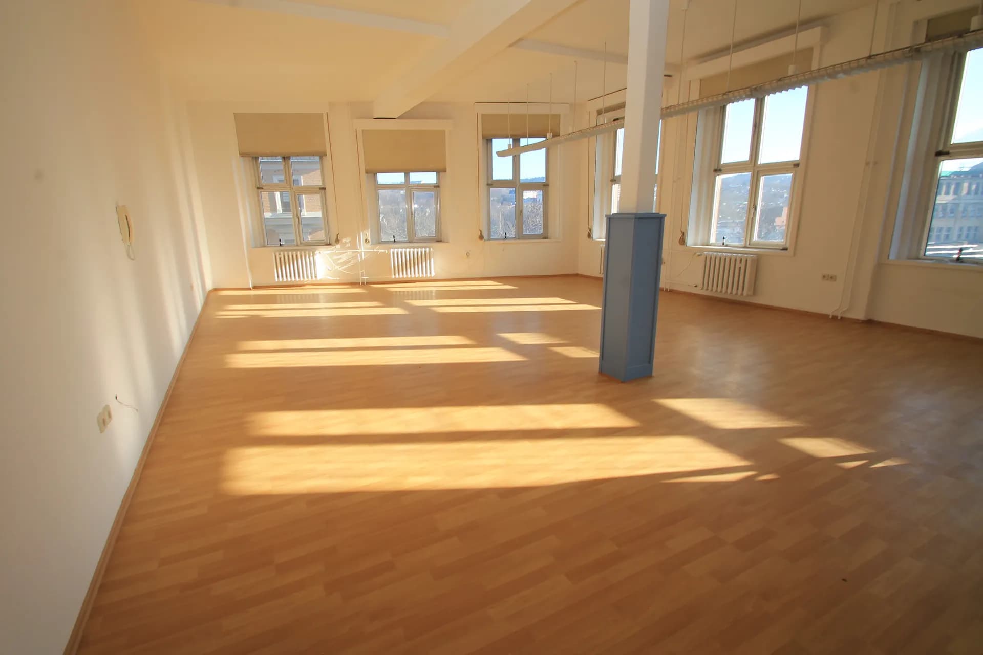Sonnendurchfluteter Großraum- oder Seminarbereich mit hellem Laminatboden, markanter Mittelsäule und durchgehender Fensterfront mit Rollos.