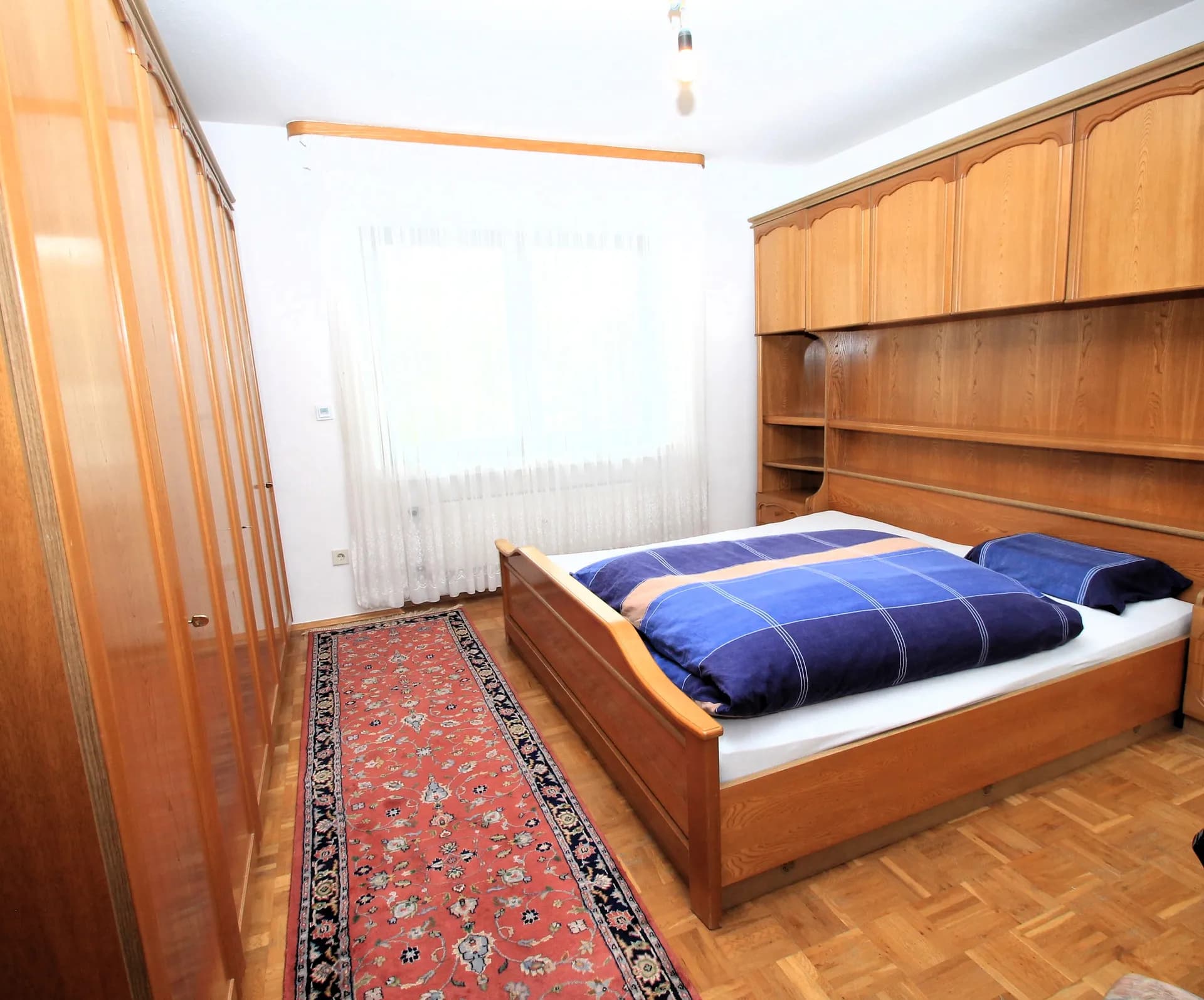 Gemütliches Schlafzimmer des Wohnhauses mit Parkettboden, Holz-Doppelbett samt integrierter Überbau-Schrankwand, großzügigem Kleiderschrank, Teppichbrücke und hellem Fenster.