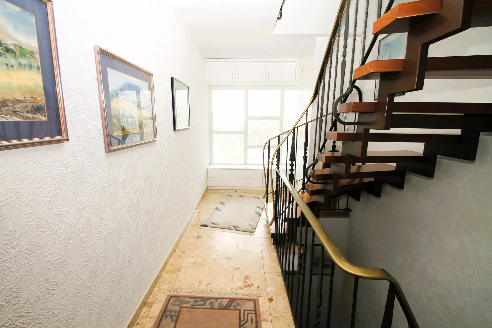 Heller Dielen- und Treppenbereich des Wohnhauses mit offener Holztreppe samt geschmiedetem Geländer, Wandbildern, hellem Fliesenboden und großem Tageslicht-Treppenhausfenster.