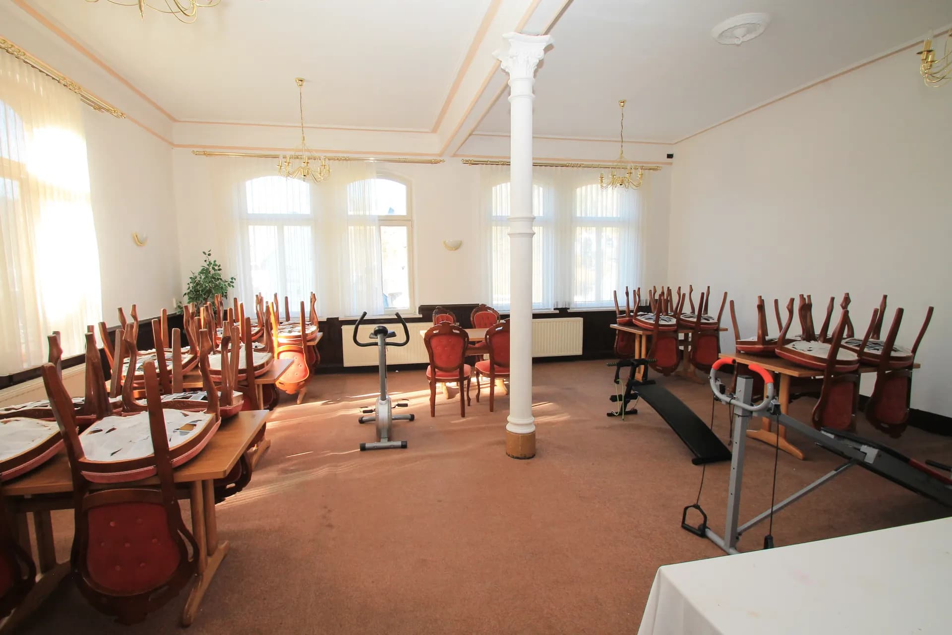 Großer multifunktionaler Saal mit Teppichboden, weißer Ziersäule, Kronleuchtern, hochgestellten Restauranttischen und aufgestellten Fitnessgeräten.