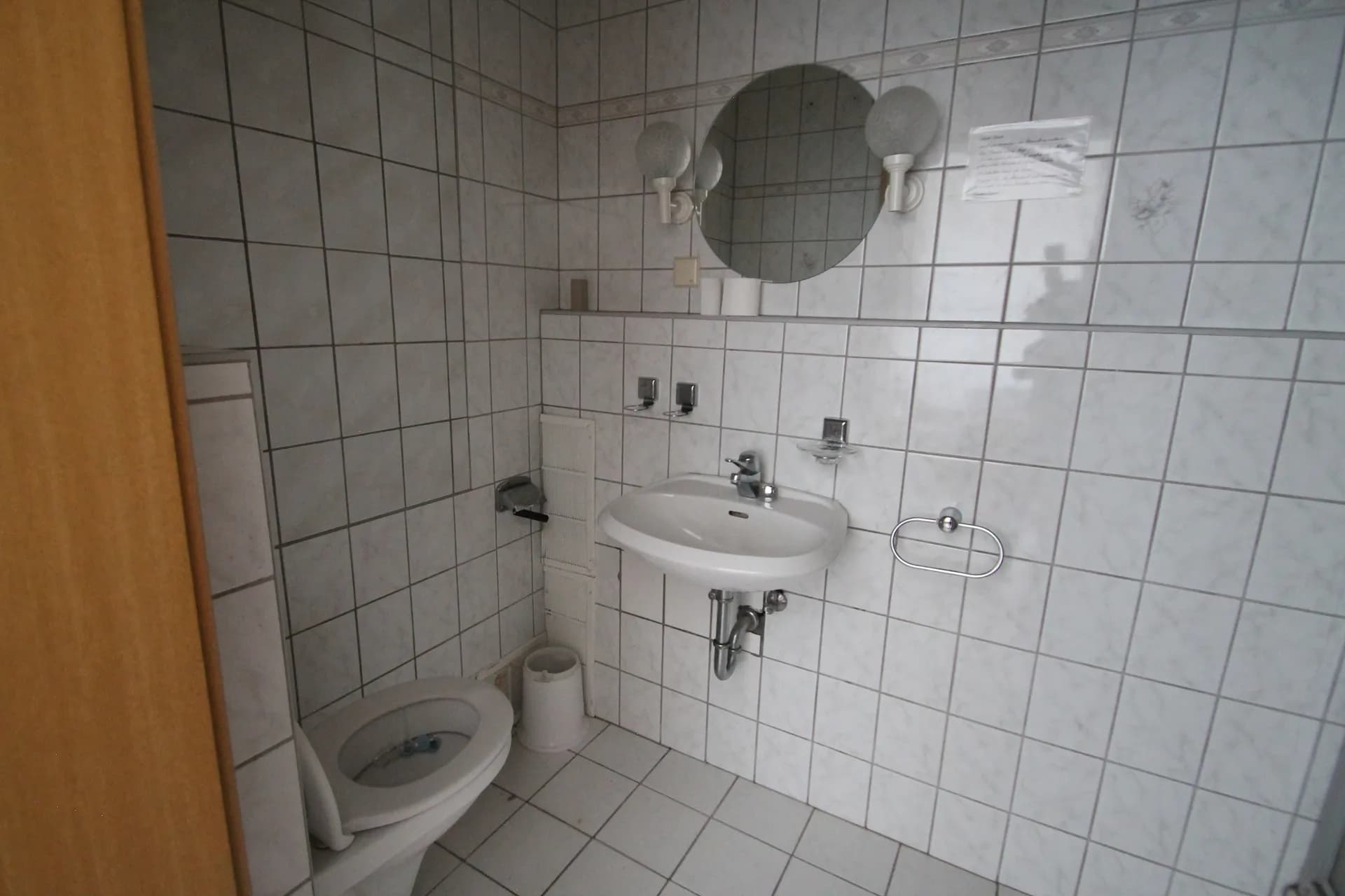Vollständig weiß gefliestes Hotel-Badezimmer mit Wandwaschbecken, rundem Spiegel samt Kugelleuchten, Armaturen und Stand-WC.