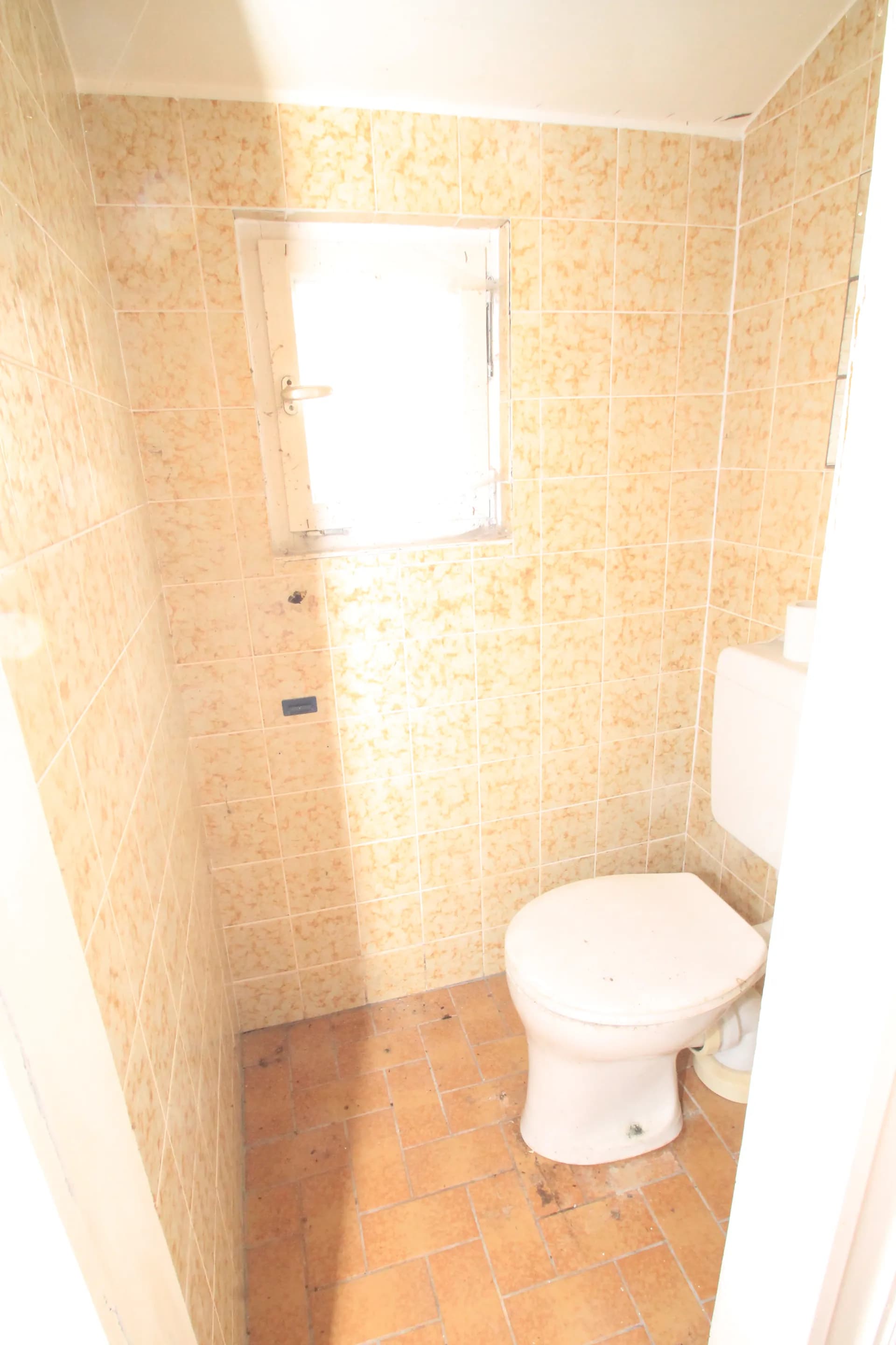Kompaktes Gäste-WC mit beigem Fliesenspiegel, Terrakotta-Bodenfliesen und kleinem Fenster zur natürlichen Belüftung.
